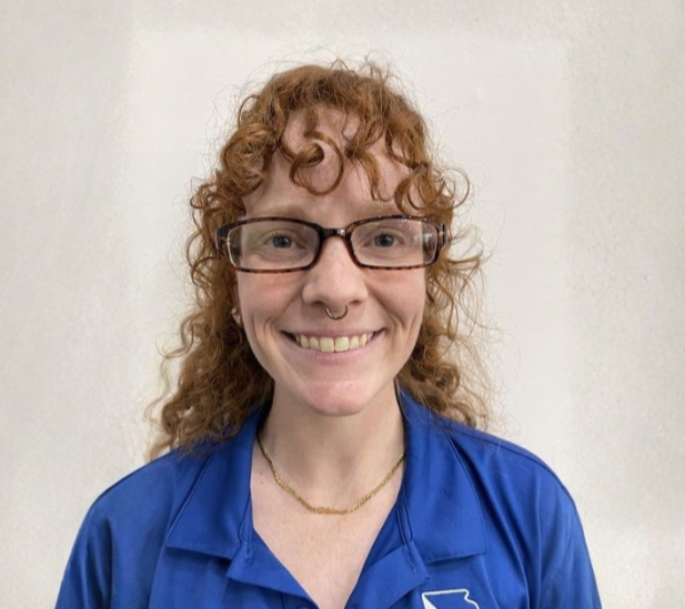 Smiling woman with curly red hair, glasses, and a septum piercing, wearing a blue polo shirt, standing against a plain wall.