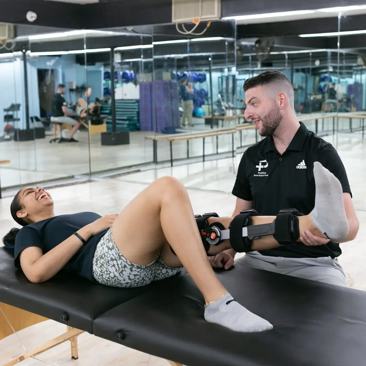 A woman lying on a treatment table, laughing, as a male physical therapist holds her leg with a robotic device at a gym or rehabilitation center.