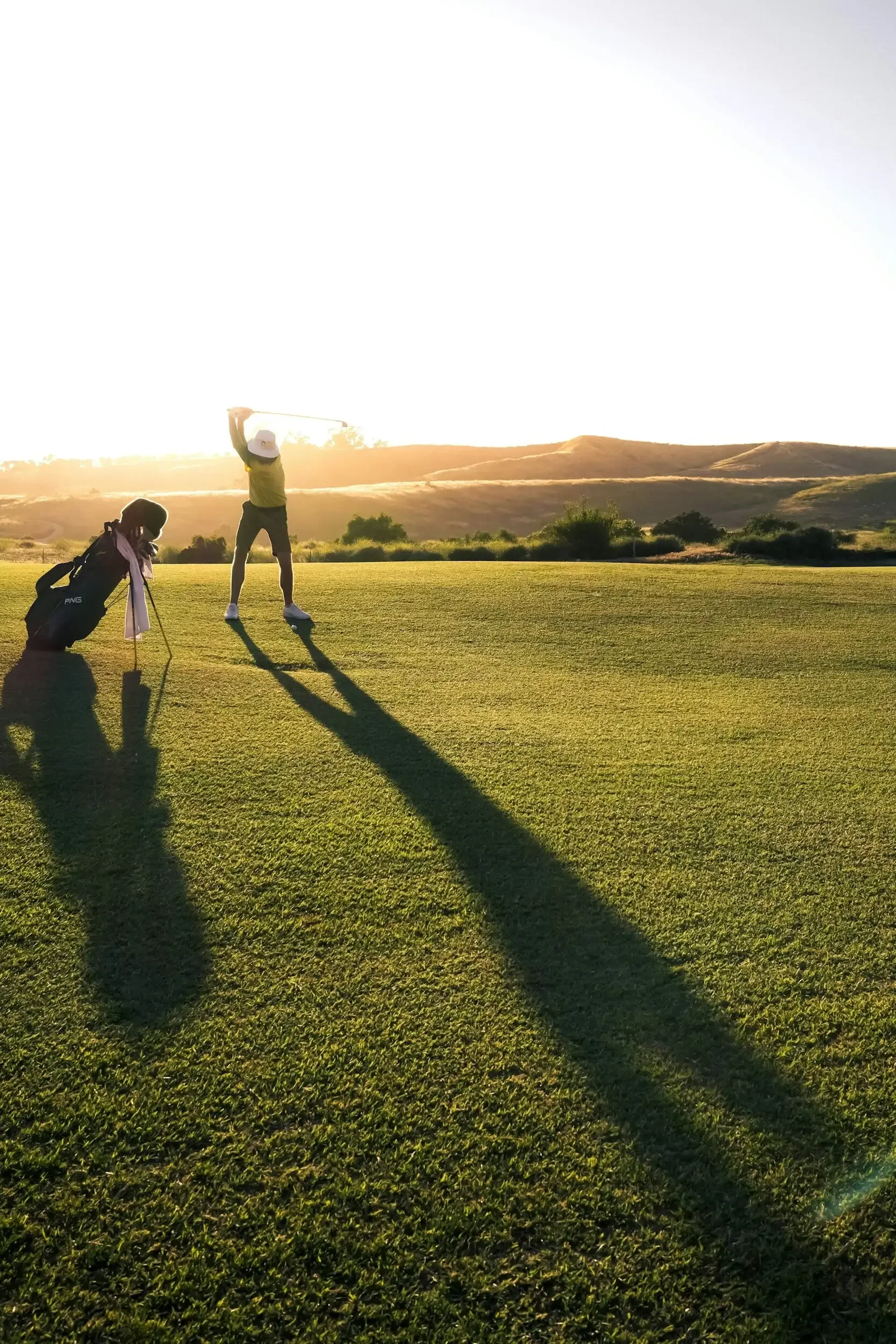 A person playing golf at sunset, swinging a club on a lush green golf course, with another person crouching nearby, and hills in the background.