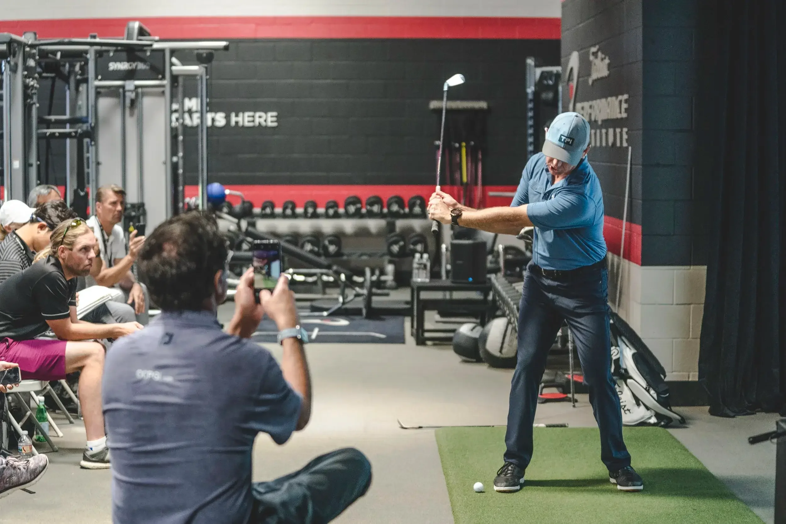A man in a blue shirt and cap practicing golf swings indoors, with a group of people seated and recording him on their phones in a gym or training facility.