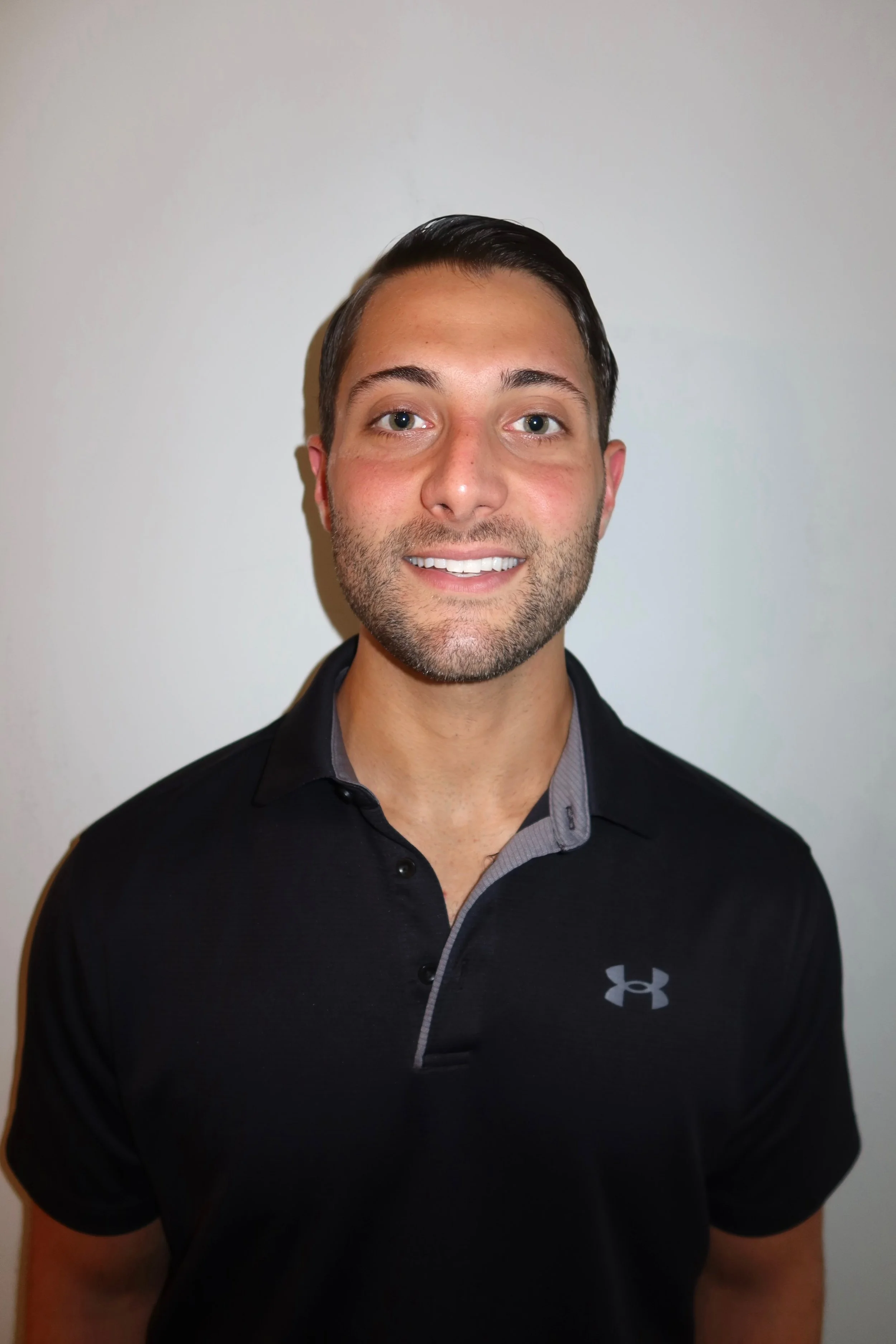 A man with short dark hair, light facial hair, and blue eyes, smiling, wearing a black Under Armour polo shirt with a gray inner collar, standing against a plain light-colored wall.
