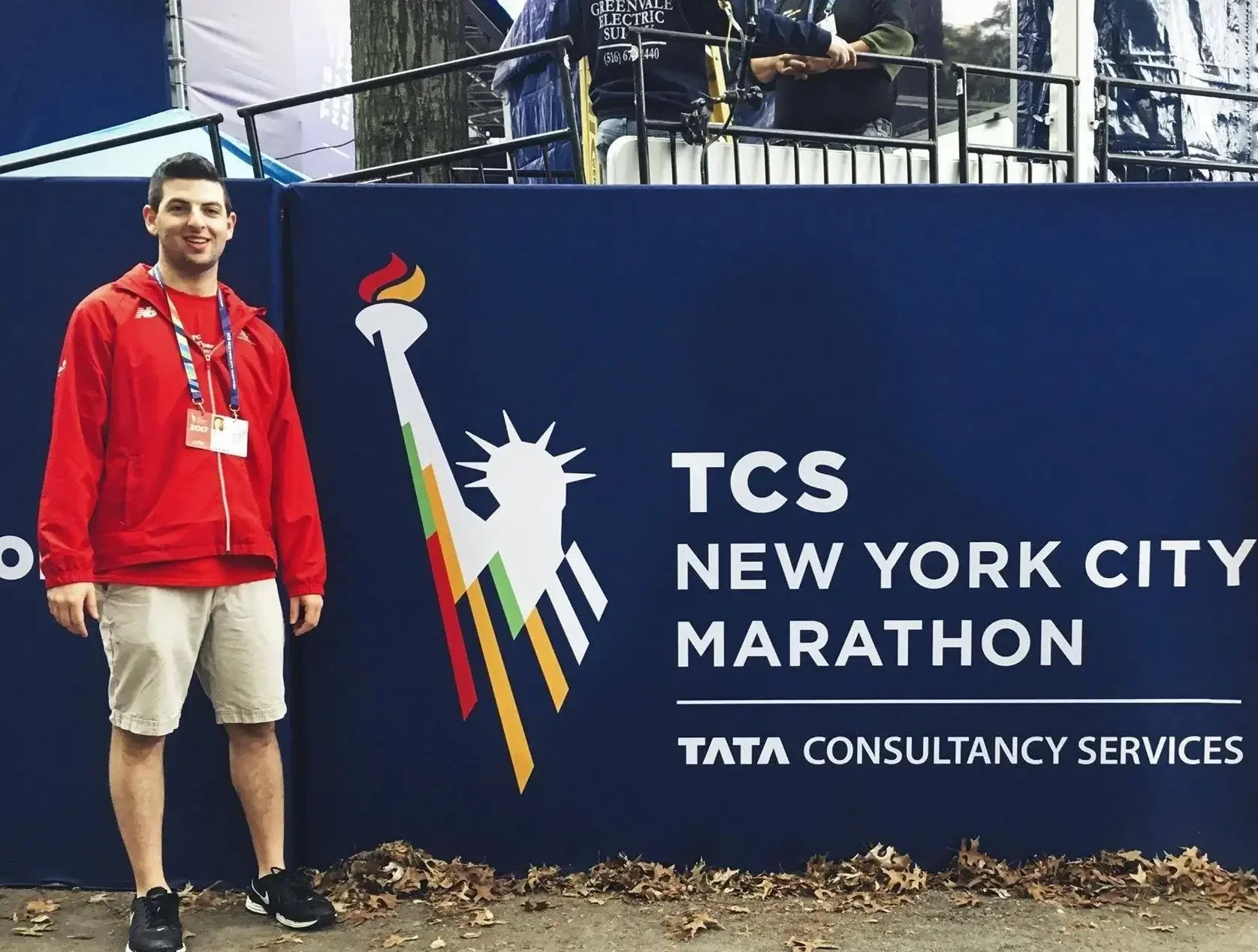 A young man in a red jacket and beige shorts standing next to a sign for the TCS New York City Marathon, with the Statue of Liberty logo, at the marathon event.