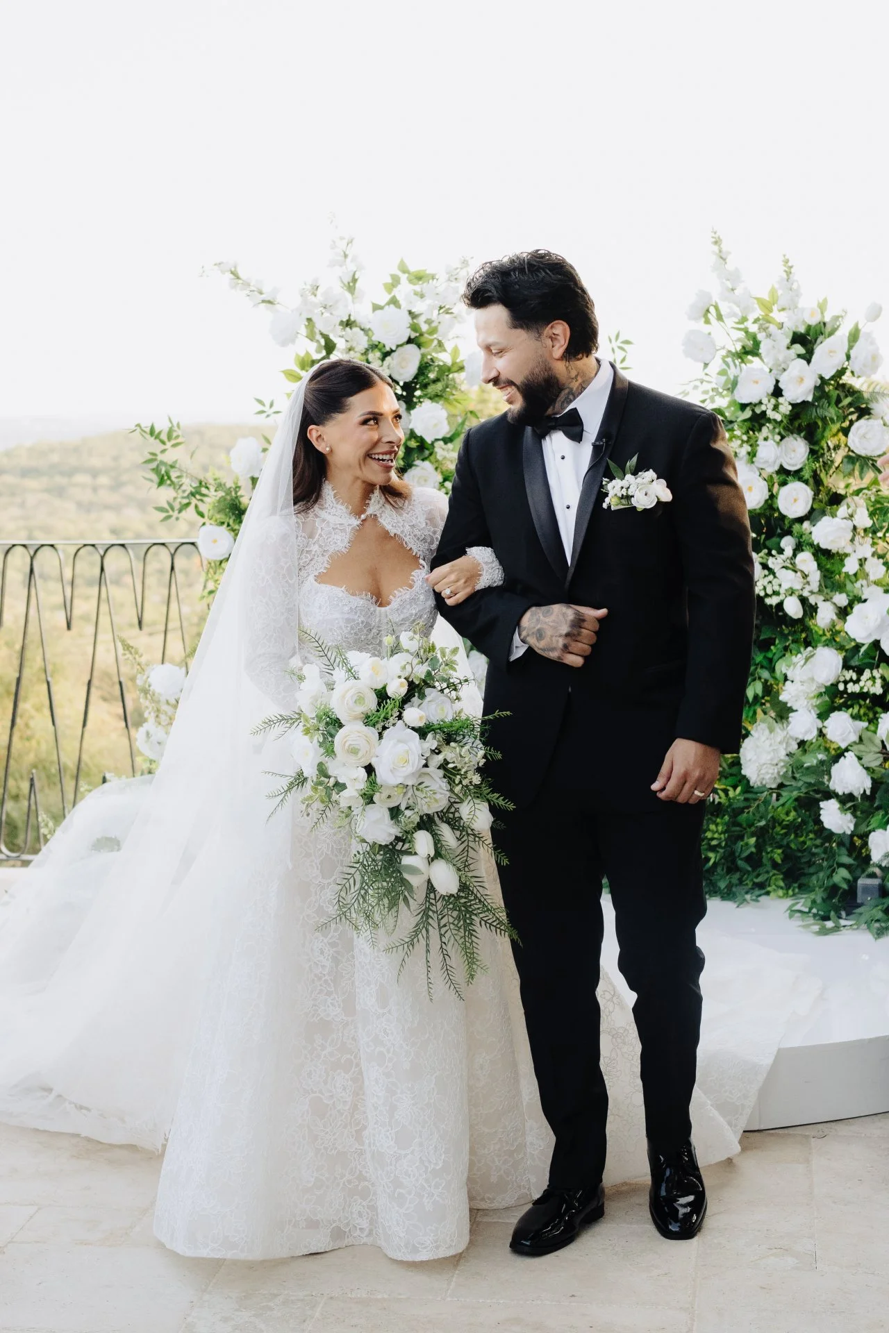 A bride and groom smiling at each other during their wedding ceremony, with a backdrop of white flowers and greenery on a balcony.