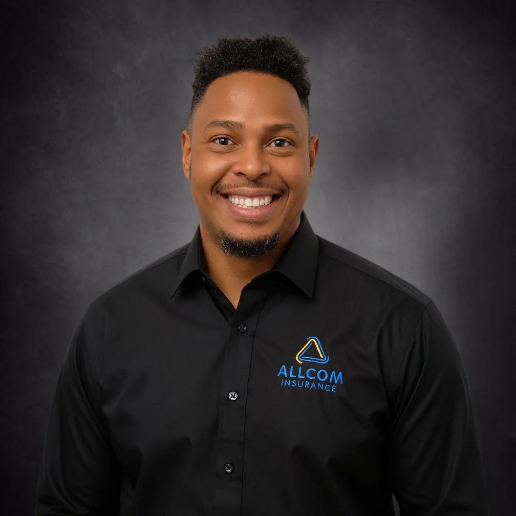 A smiling man with short curly hair and a goatee wearing a black polo shirt with a logo that reads Allcom Insurance.