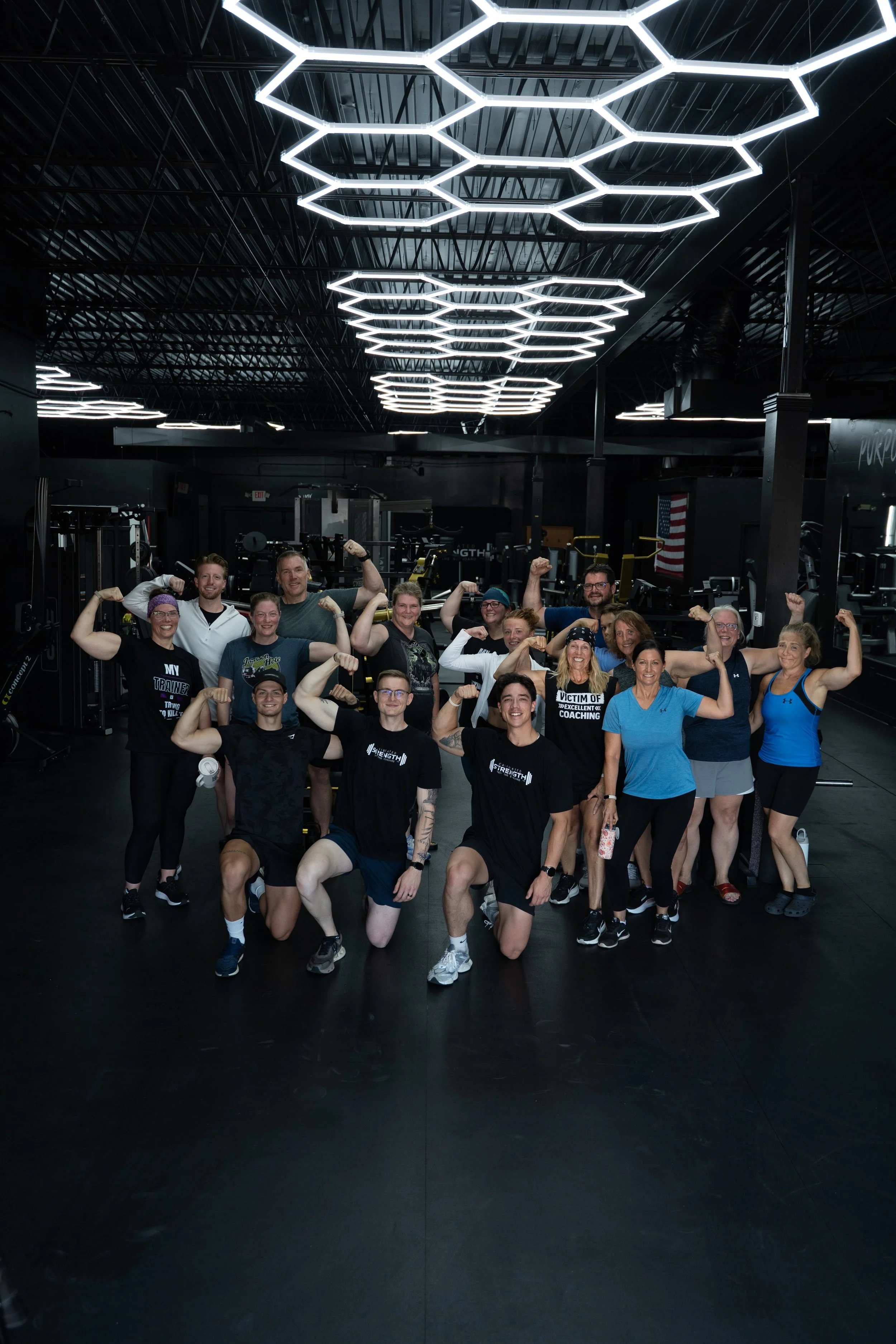 Group of people in a gym,posed in front of exercise equipment, all flexing their arm muscles.