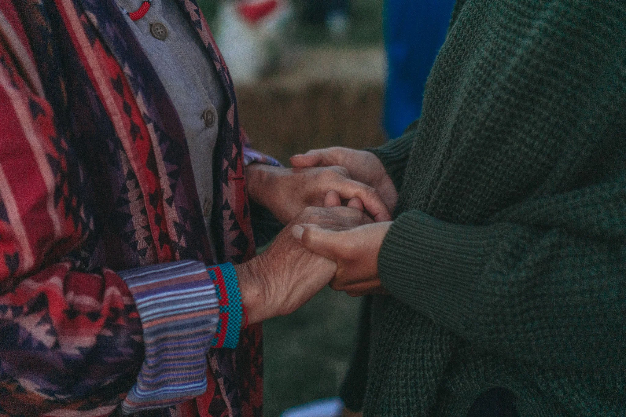 Two people holding hands, one appears to be older and wearing a colorful patterned jacket, the other is younger wearing a dark green sweater.