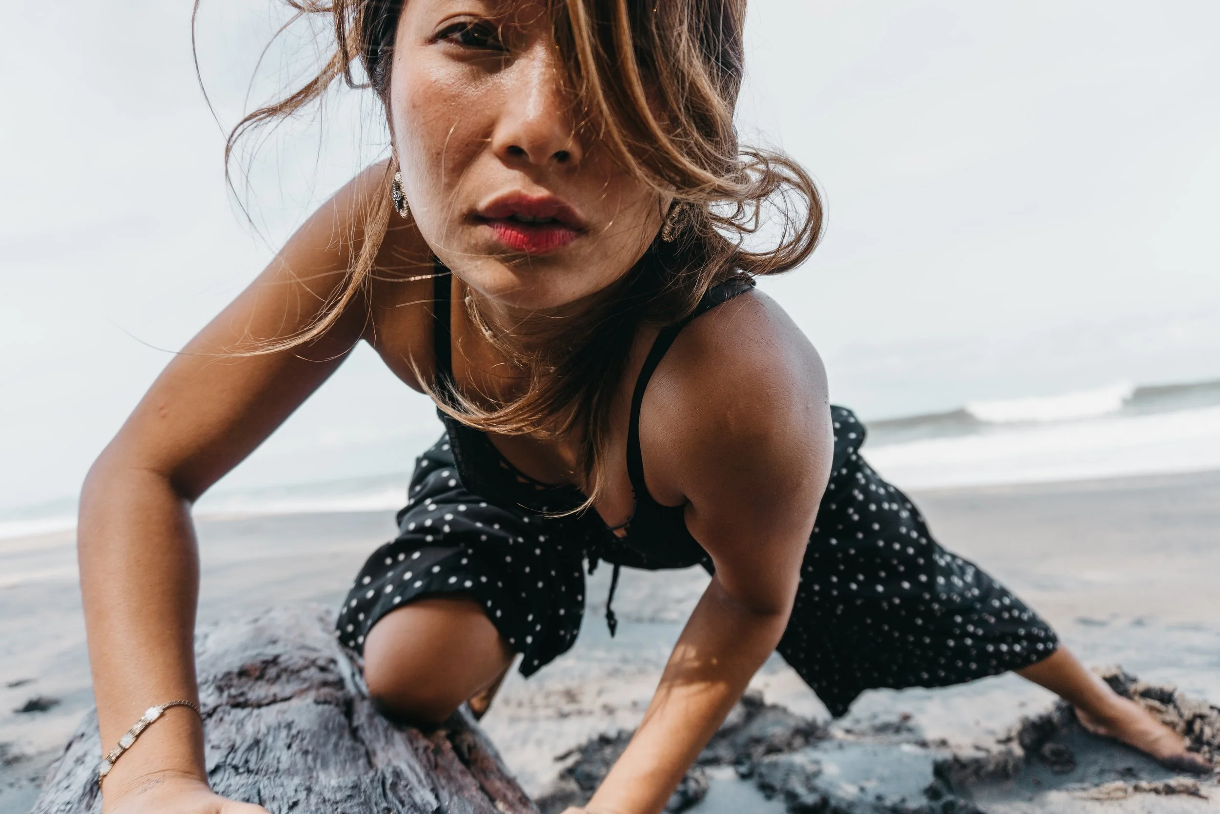 Woman in black polka dot dress crouching on a beach with waves in the background.