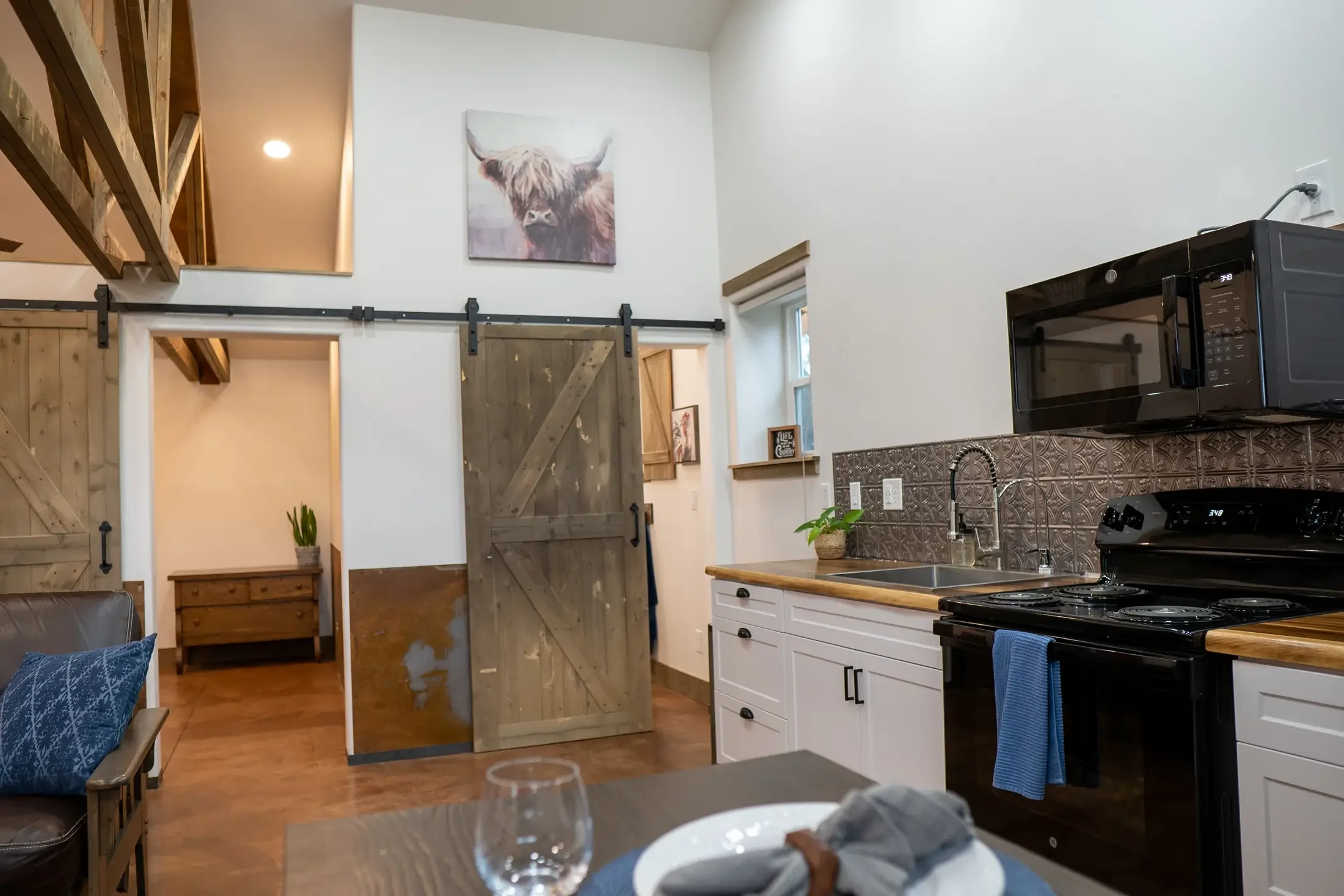 Interior of a modern farmhouse-style kitchen with white cabinets, a black microwave, and decorative patterned backsplash. A rustic sliding barn door leads to a hallway, and a high ceiling features a wall art of a Highland cow. There are plants on the
