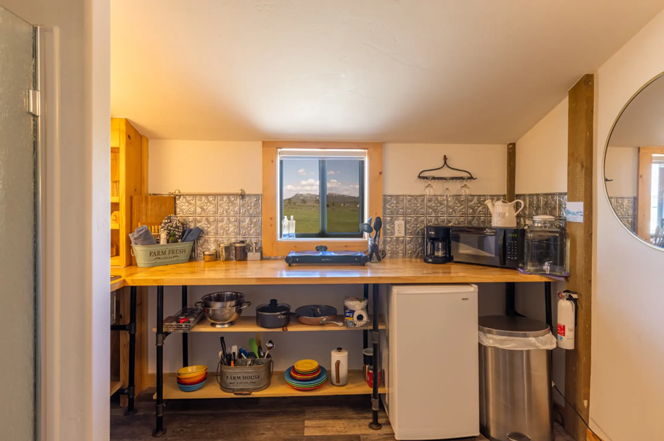 View of a rustic kitchen with natural wood accents, a window overlooking a green landscape, and various kitchenware on open shelves and countertops.