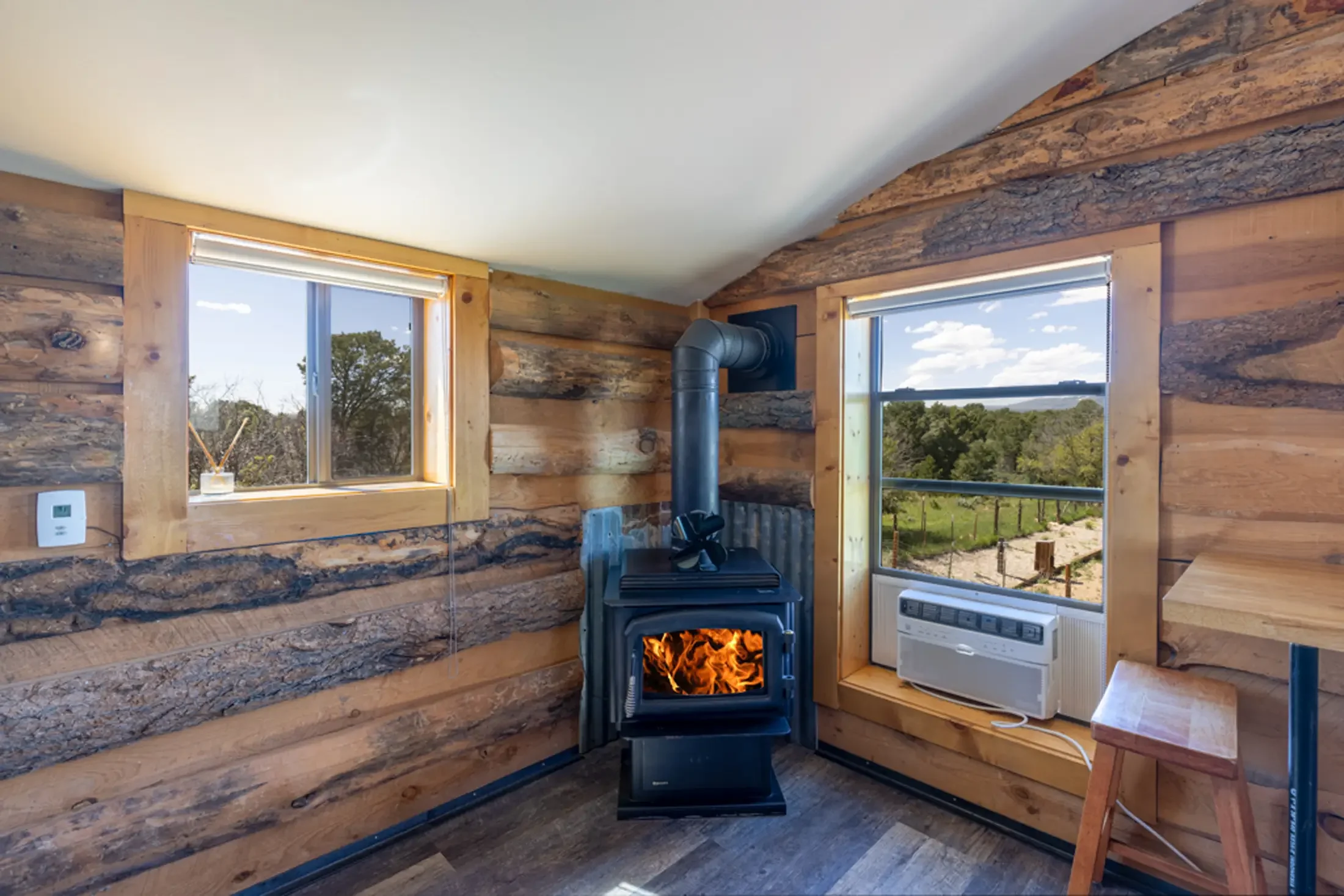 Interior of a rustic wooden cabin with large windows, a wood stove emitting flames, a small dresser with a white heater, and outdoor scenery of trees and sky visible through the windows.