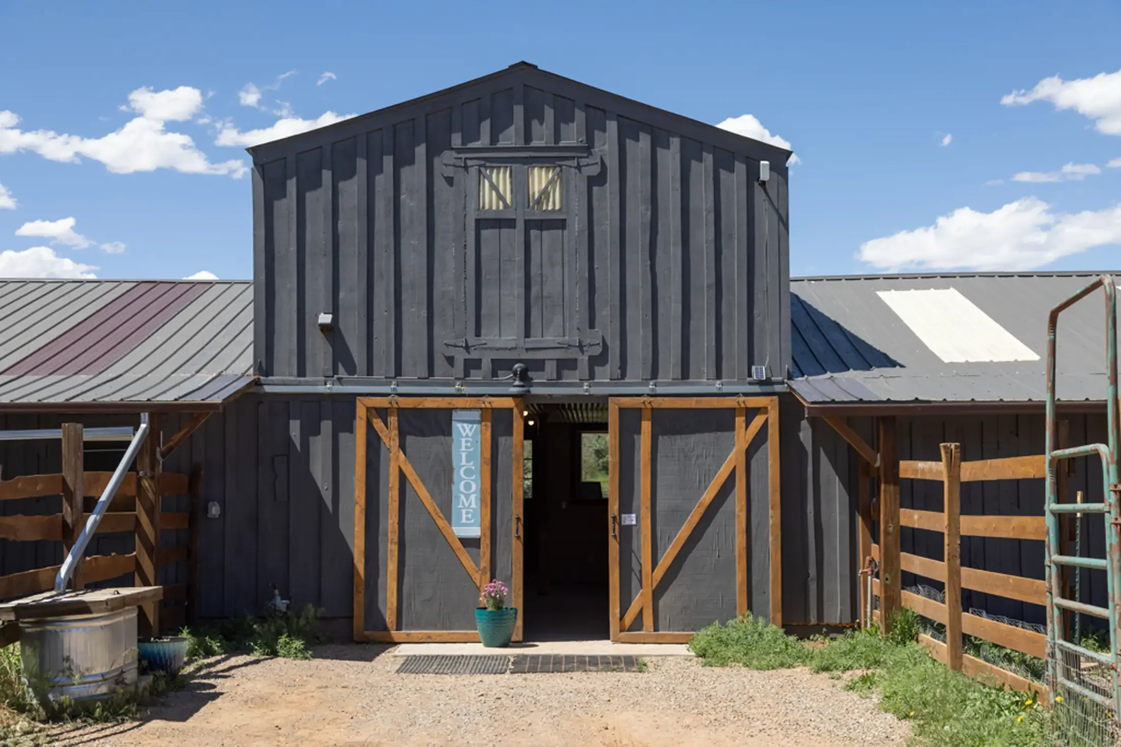 A rustic barn with gray vertical wood siding, a small window with yellow curtains, and a wooden gate with a wheat design. The barn has a metal roof and is surrounded by some plants, with a blue sky and clouds in the background.