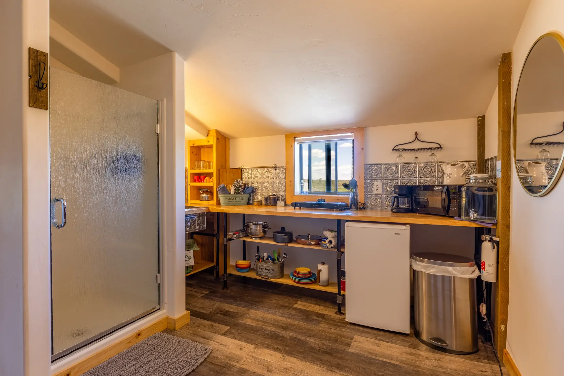 View of a cozy kitchen with a wooden countertop, open shelves, a small refrigerator, and a coffee maker, with a window showing a landscape outside.