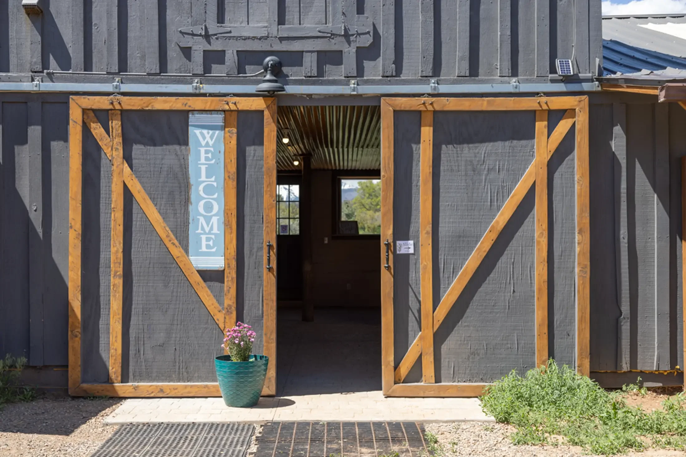 The image shows the front of a barn conversion with black painted wood and a large, open doorway framed in fresh, light wood. There is a welcome sign next to the door and a potted plant with pink flowers in front. Small plants and grass are around th