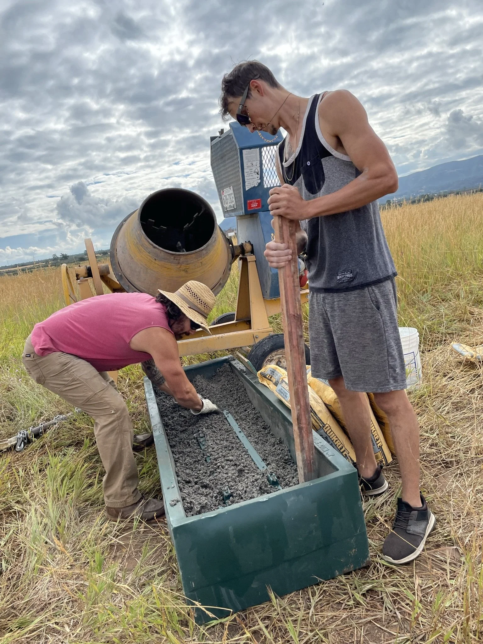 Two men are working outdoors, pouring concrete into a mold, with one using a metal tool and the other operating a cement mixer, in a field with tall grass and mountains in the distance under a cloudy sky.