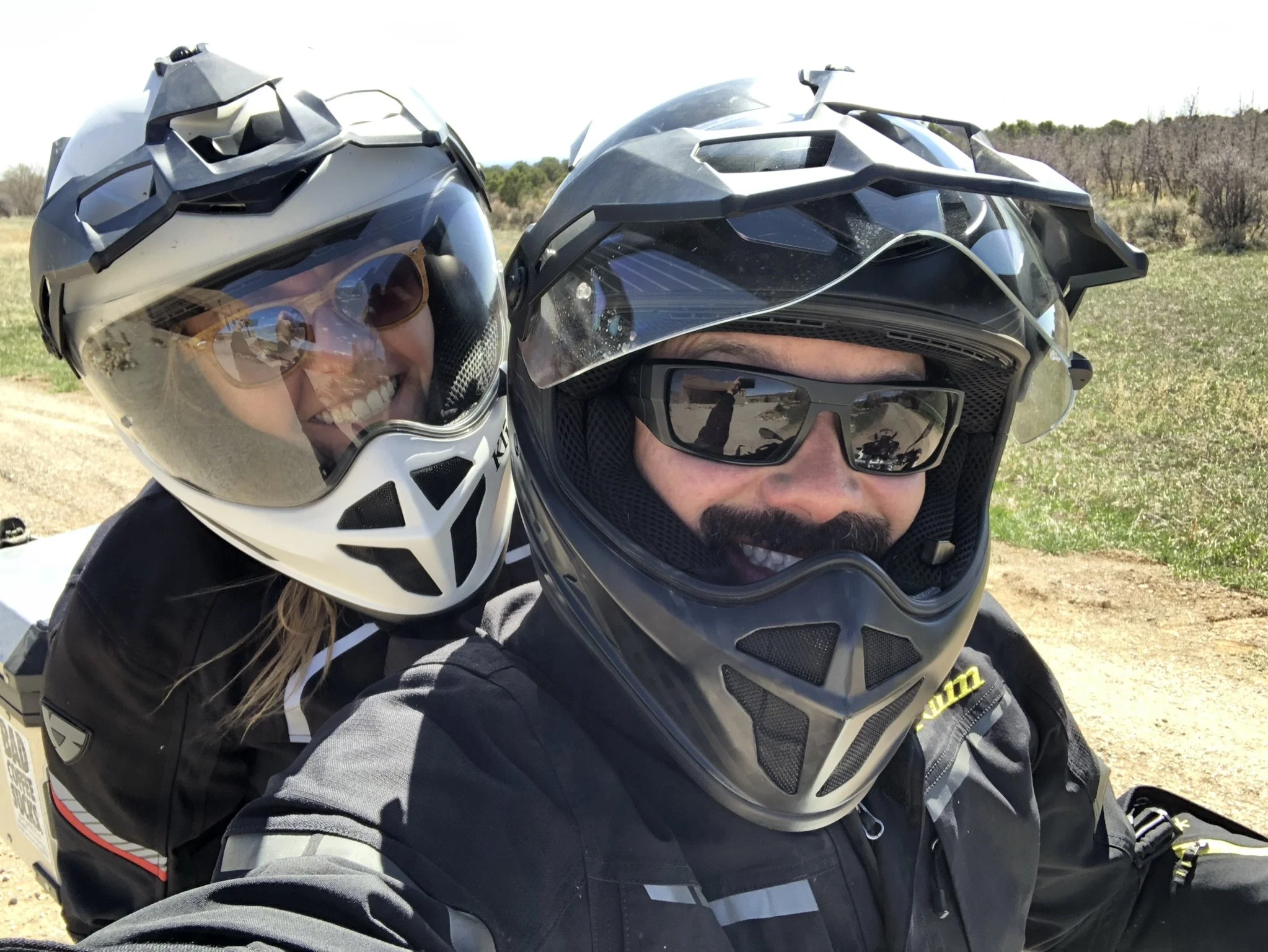 Two people wearing motorcycle helmets, smiling, on a motorcycle outdoors in a sunny, rural landscape.