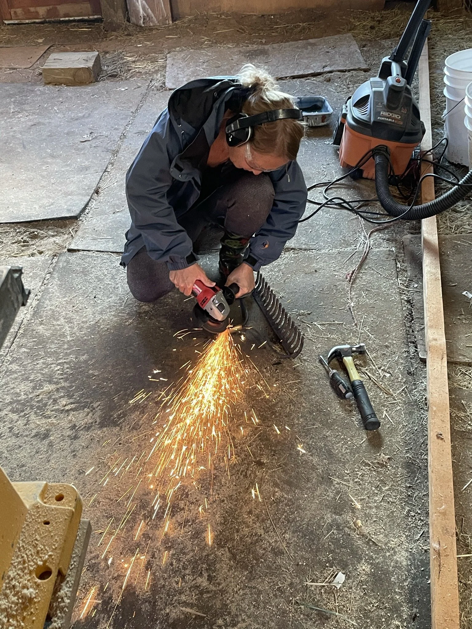 A person wearing a black jacket, hoodie, and headphones is kneeling and using a power tool to cut a metal object, creating sparks in a workshop or garage with various tools and construction materials around.