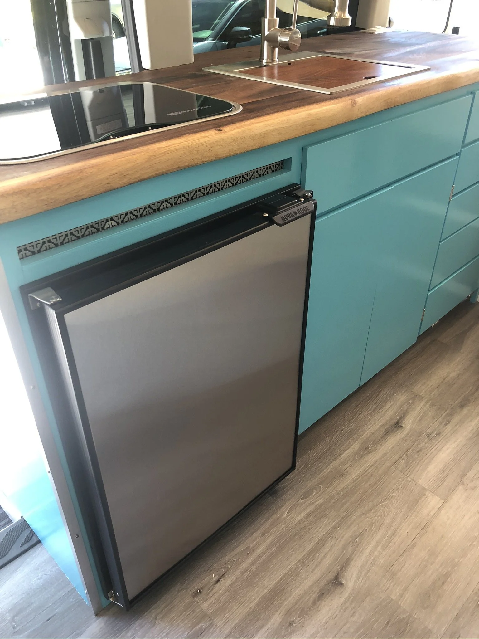 A modern kitchen with a wooden countertop, teal cabinets, a built-in stainless steel refrigerator, and a black stovetop. A stainless steel sink with a chrome faucet is also visible.