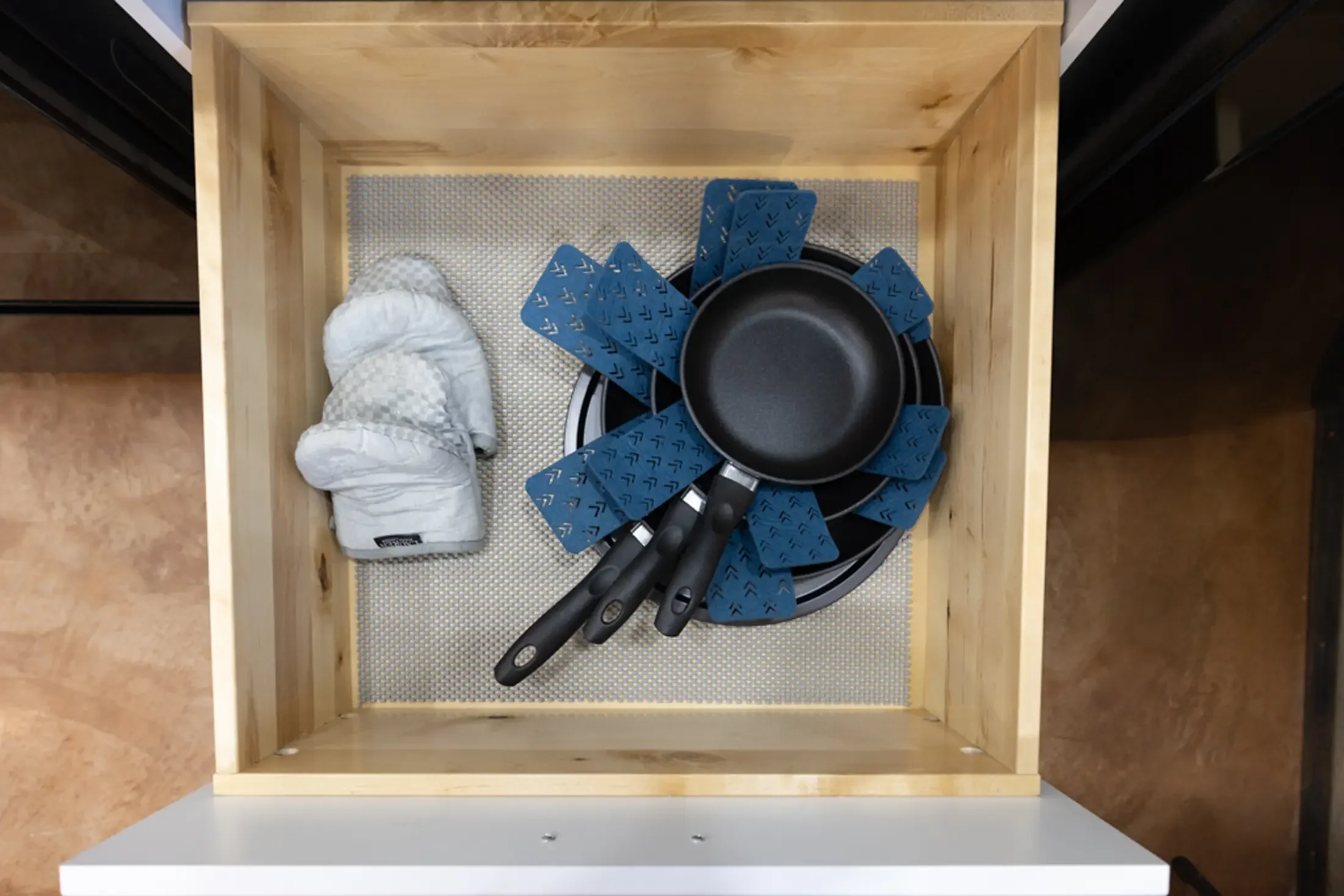 A wooden drawer contains disposable diapers, a black frying pan, blue pot holders, and multiple black-handled kitchen utensils.