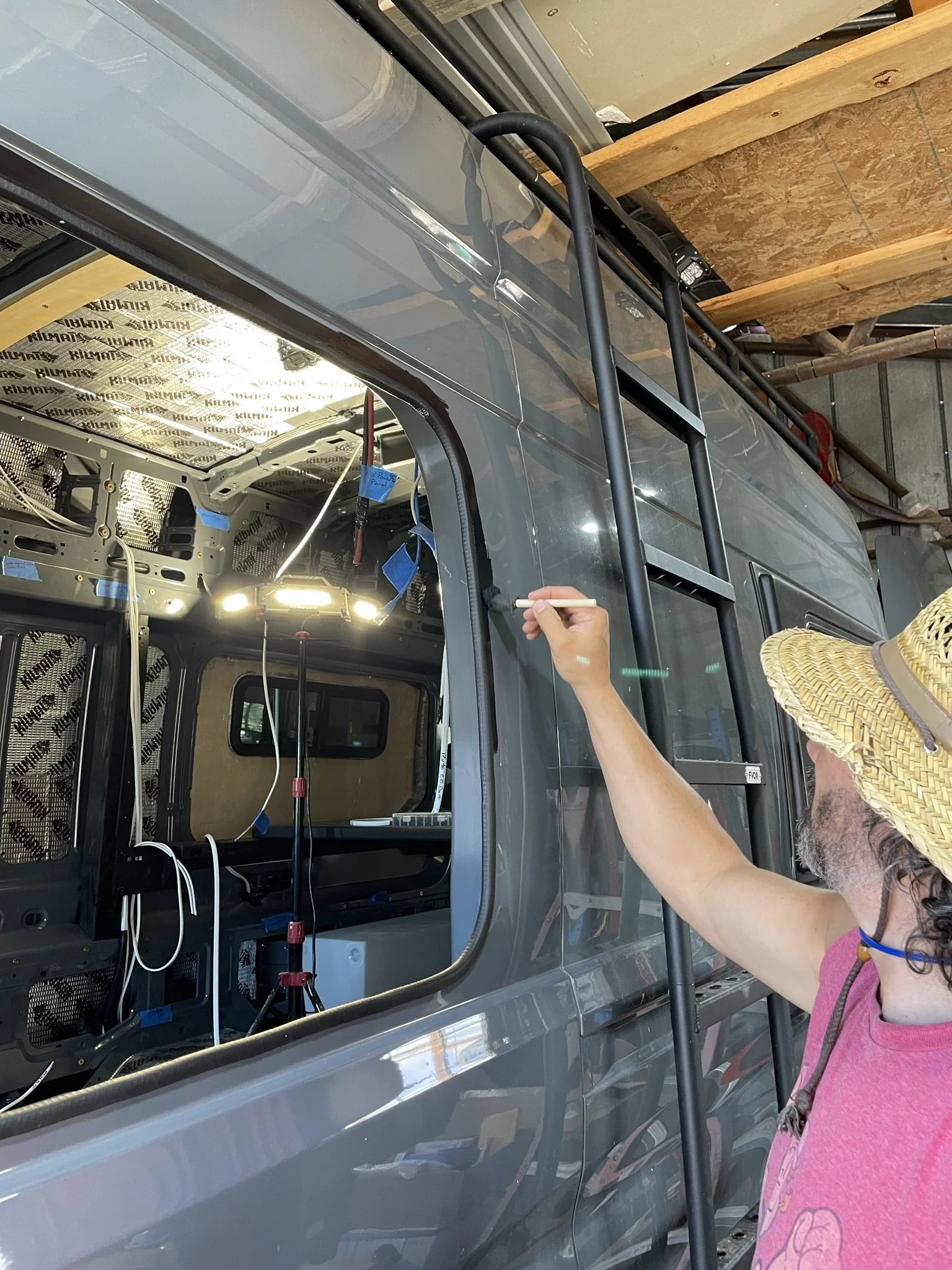 A woman wearing a straw hat and a pink shirt working on the exterior of a gray vehicle in a garage or workshop.