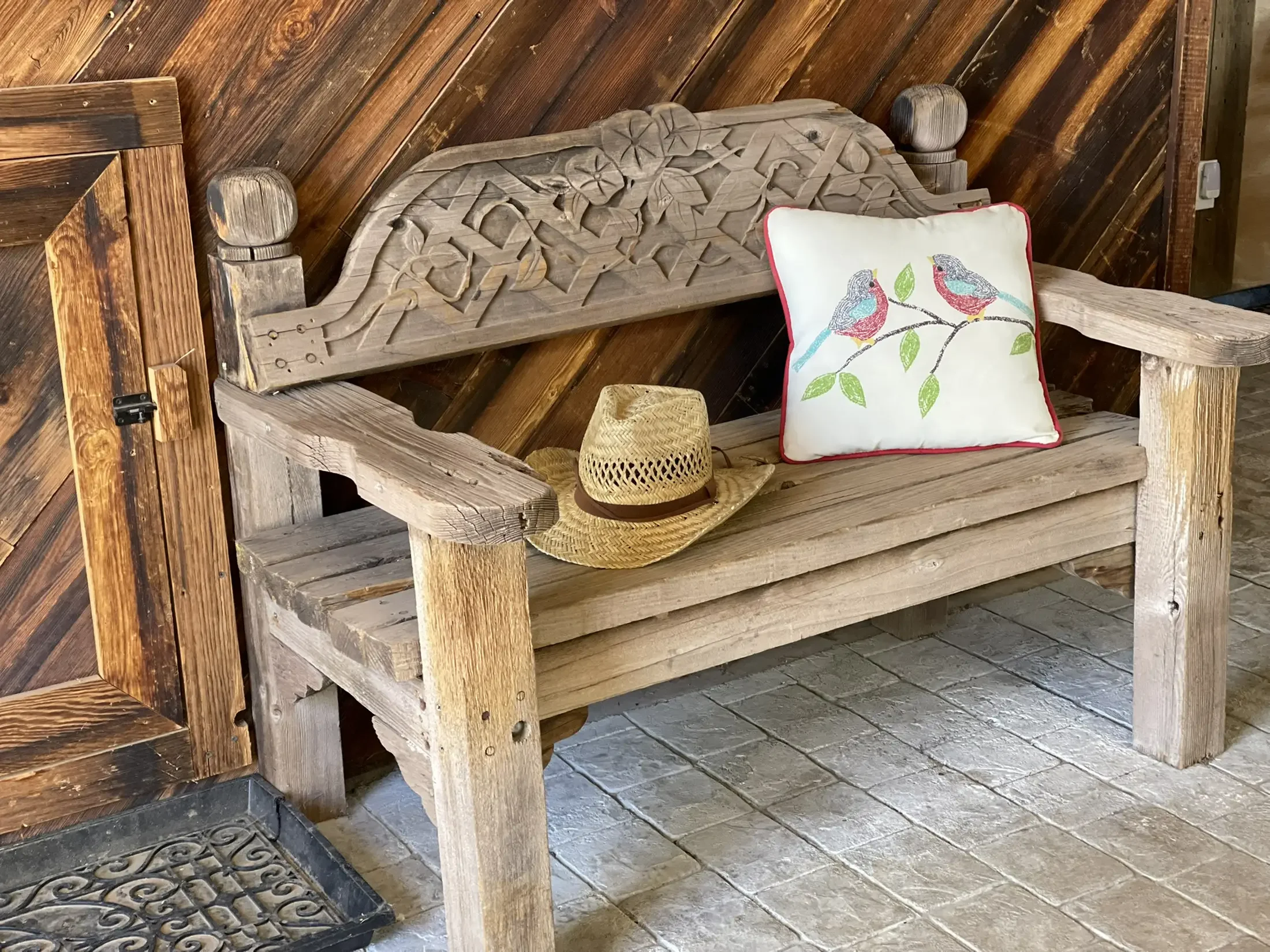 A rustic wooden bench with a decorative backrest featuring intricate carvings. A white cushion with colorful bird and leaf embroidery and red piping is placed on the bench. A straw hat with a black band rests on the bench in front of the cushion. The