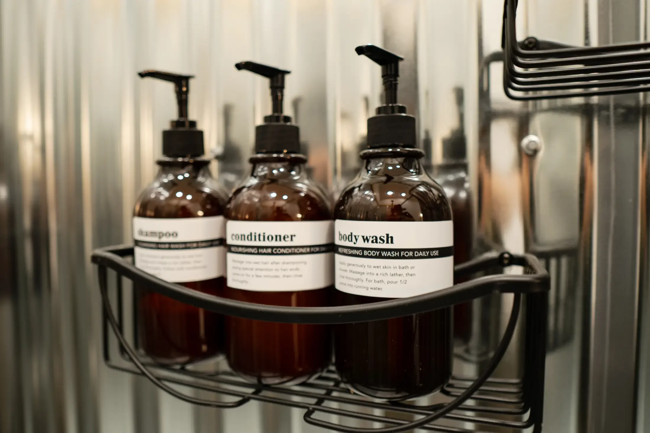 Three brown bottles with black pump dispensers labeled shampoo, conditioner, and body wash on a metal shelf in a shower or bathroom setting.