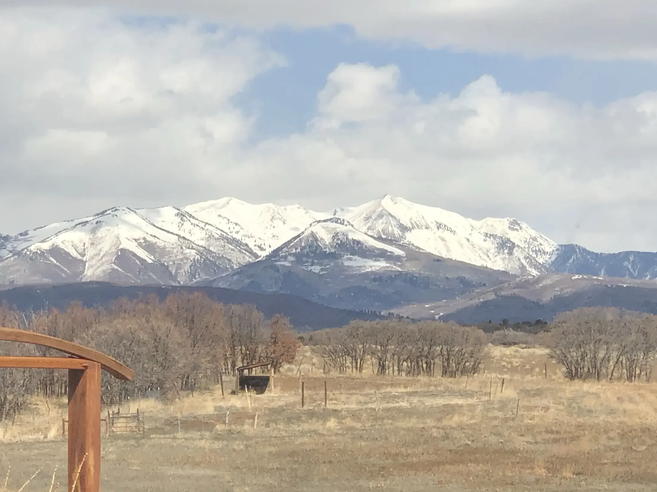 Landscape photo of snow-capped mountains under a partly cloudy sky, with trees and fields in the foreground.