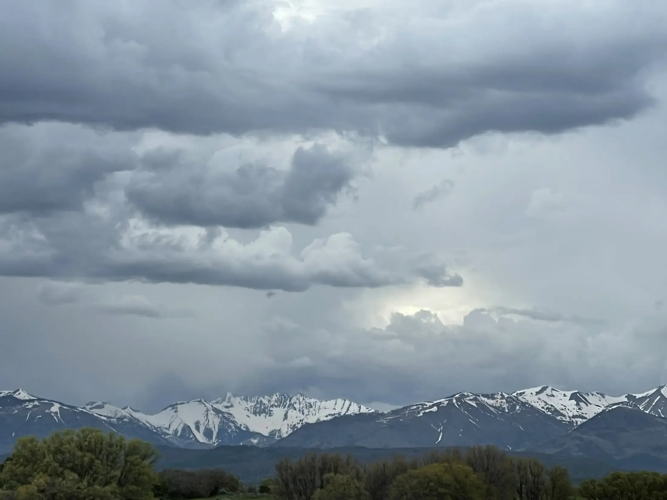Snow-capped mountains under a cloudy sky with dark clouds and some patches of light, and green trees in the foreground.