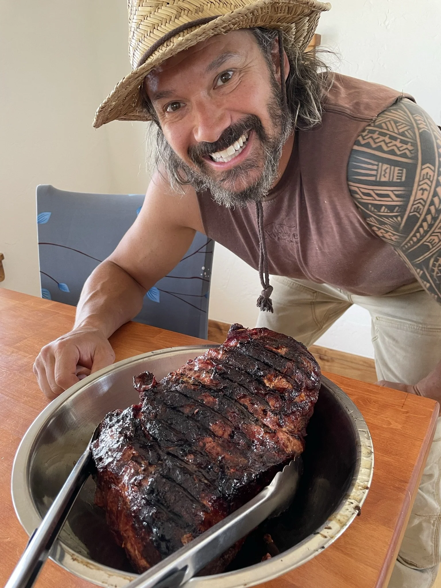 A man wearing a straw hat and a sleeveless shirt is smiling and leaning over a table with a large, cooked piece of meat in a metal tray.