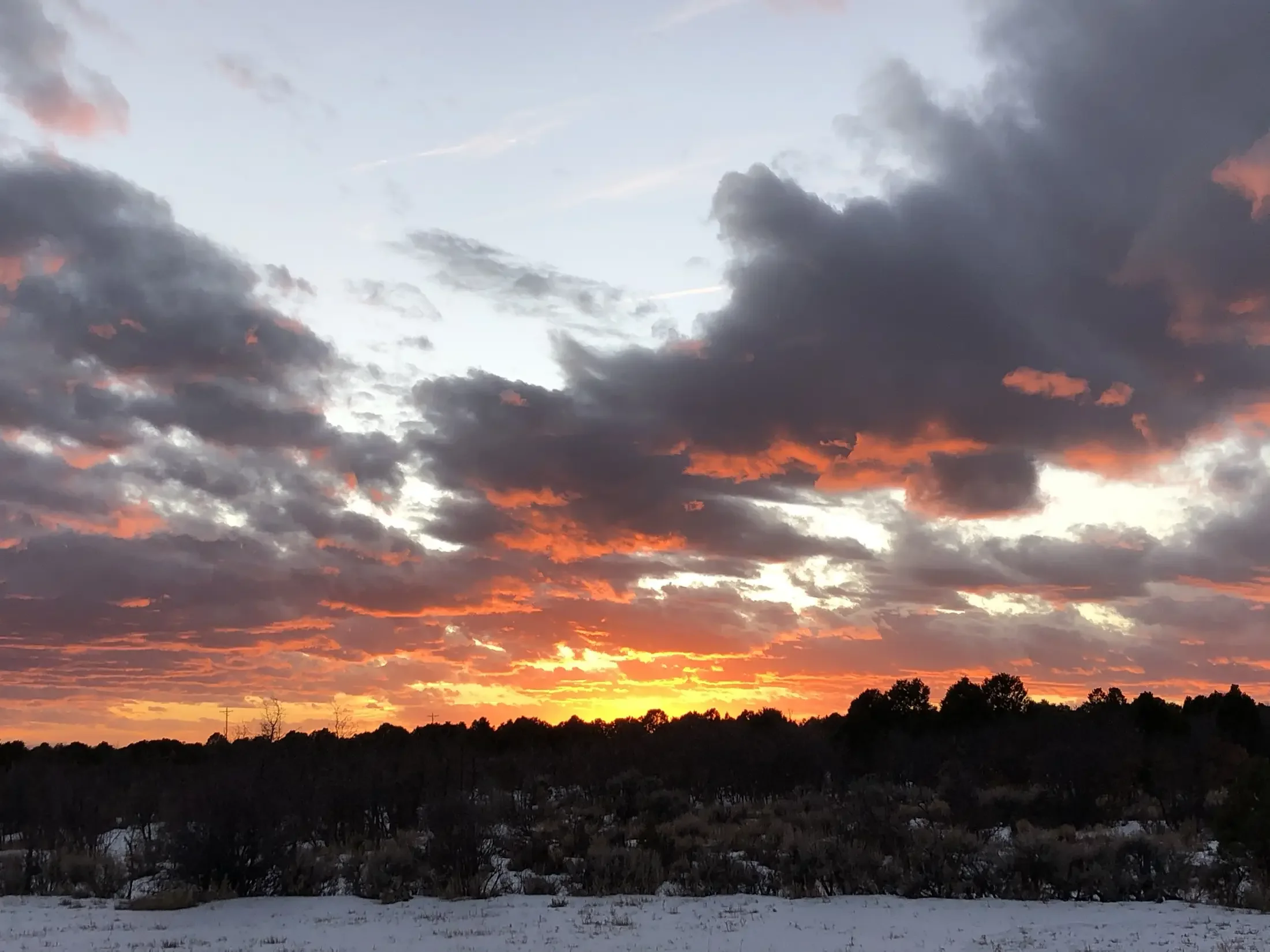 A sunset over a landscape with trees and snow-covered ground, with clouds in the sky illuminated by the setting sun.