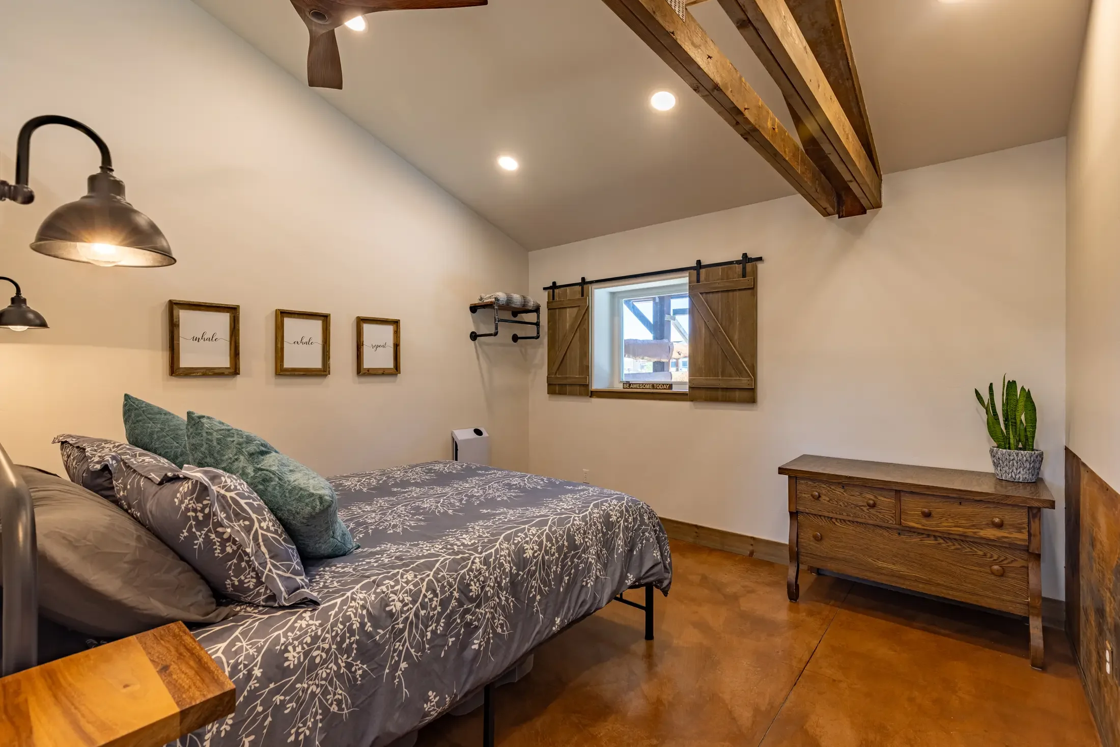 A cozy bedroom with a bed covered in gray and white floral bedding and decorative pillows, wood accents including a window shutter and a dresser, wall art, a potted plant, and rustic wood beams on the ceiling.