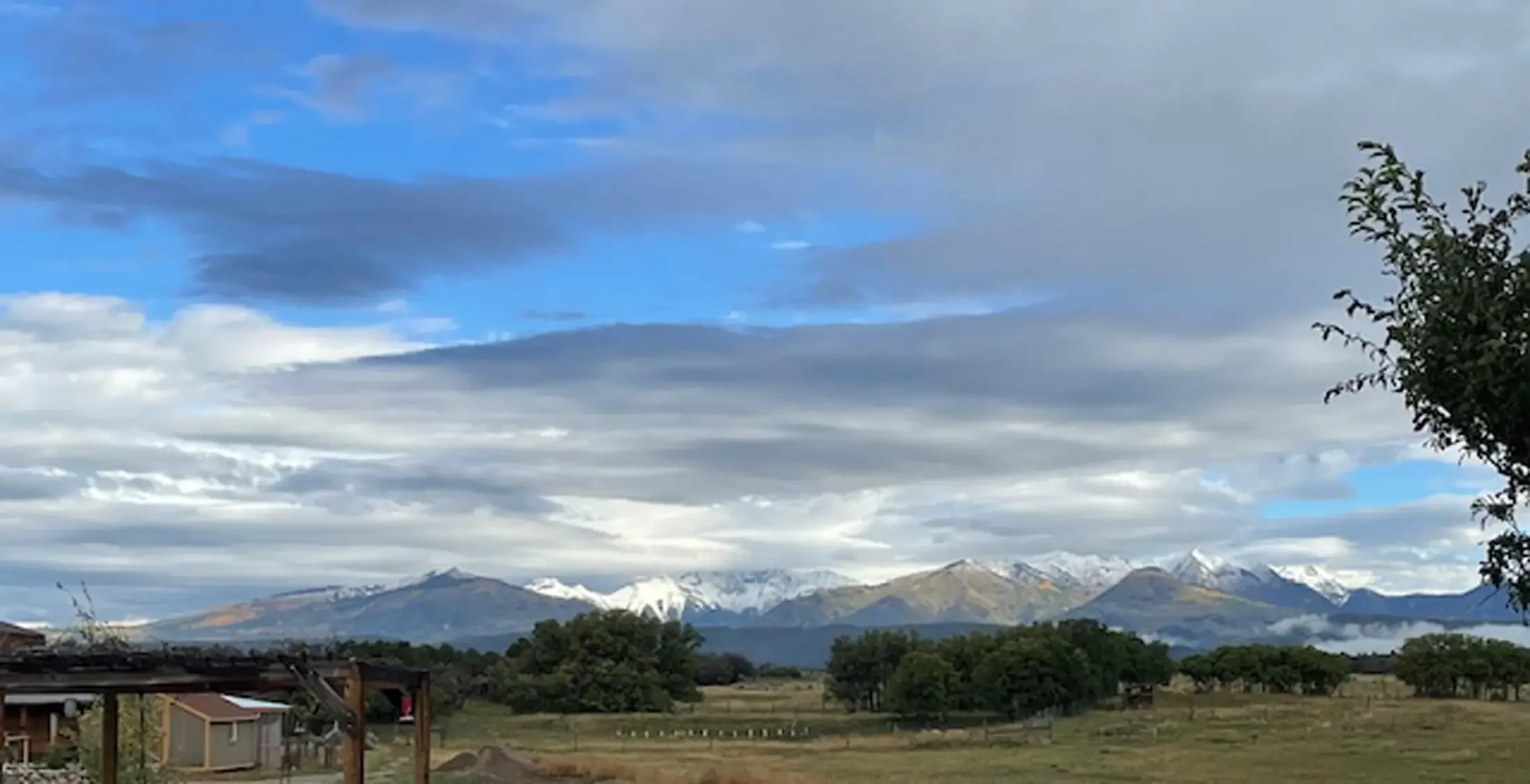 View of a mountain range with snow-capped peaks under a partly cloudy sky, with some trees and a few houses in the foreground.