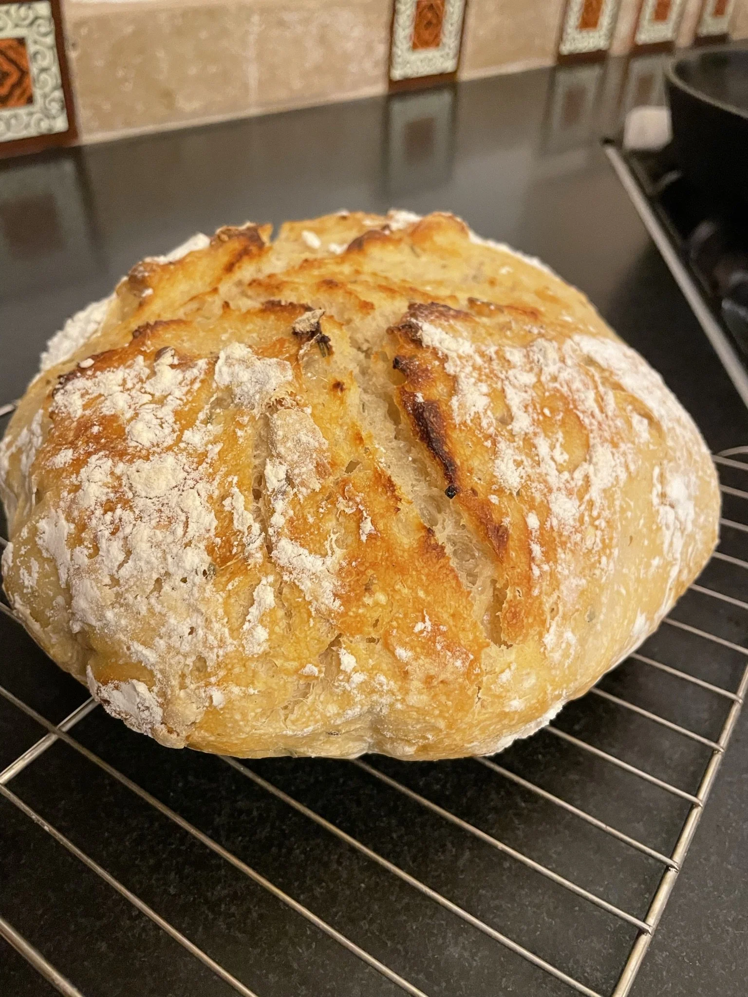 A round, rustic loaf of bread with a golden crust, dusted with flour, resting on a wire cooling rack in a kitchen.