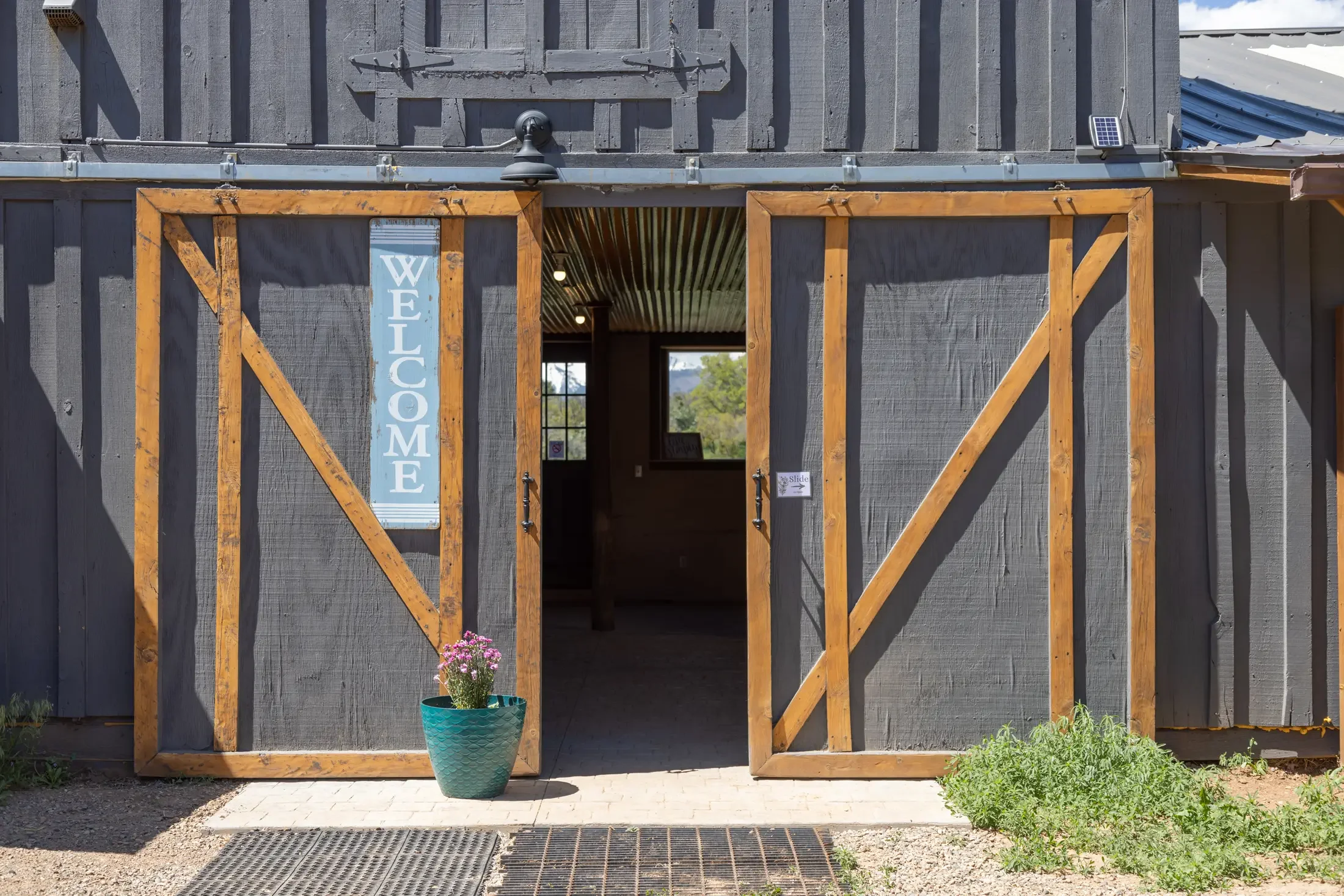 Exterior of a barn with gray wooden siding and large sliding barn doors framed with natural wood. The left door is open, revealing an interior with a window showing trees and mountains outside. A tall, blue flower pot with pink flowers is placed in f