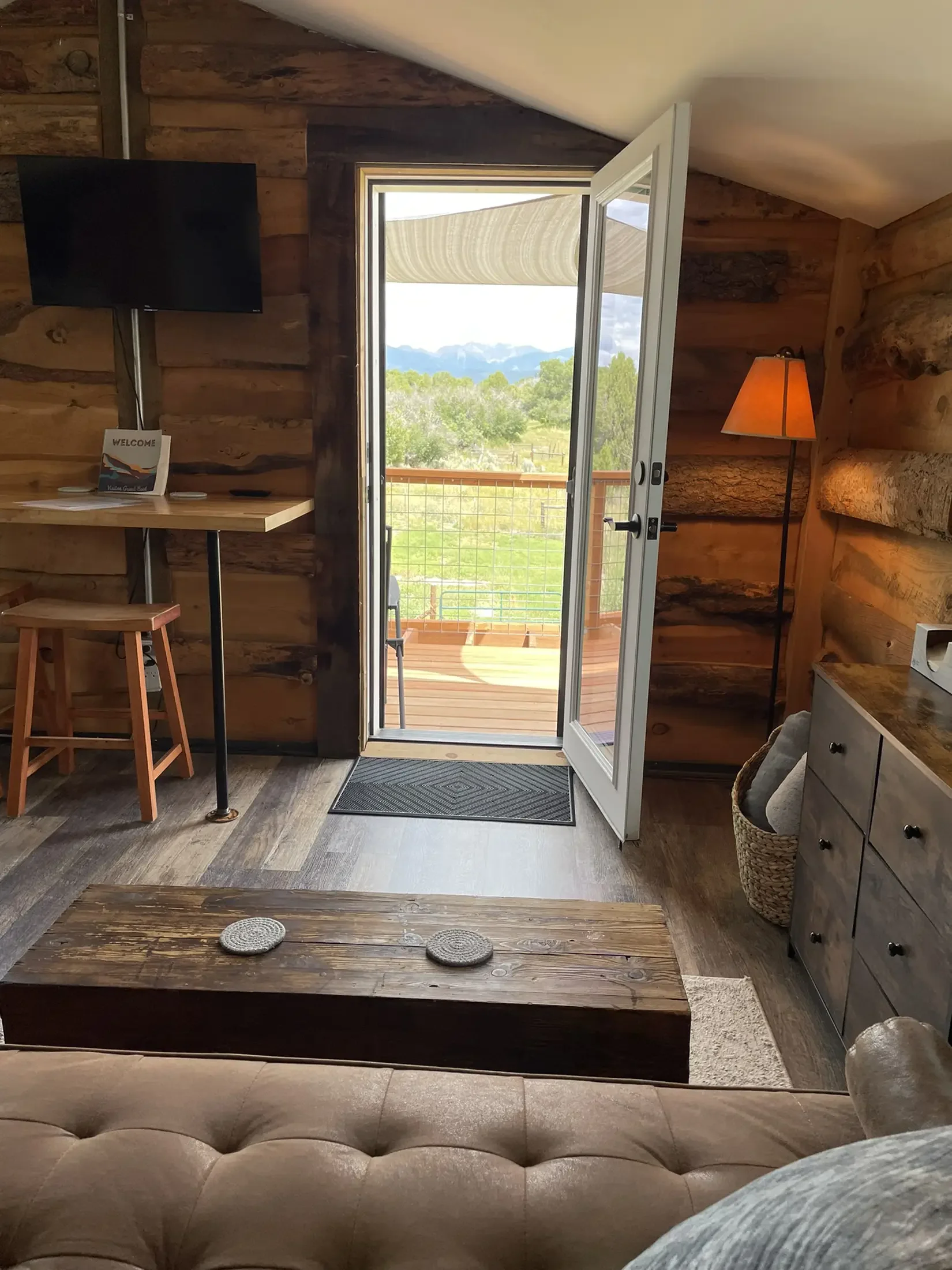 Interior view of a rustic cabin with wooden walls, a small balcony visible through a door, a TV mounted on the wall, a lamp, a wicker basket, a dresser, and a sofa with a pillow.