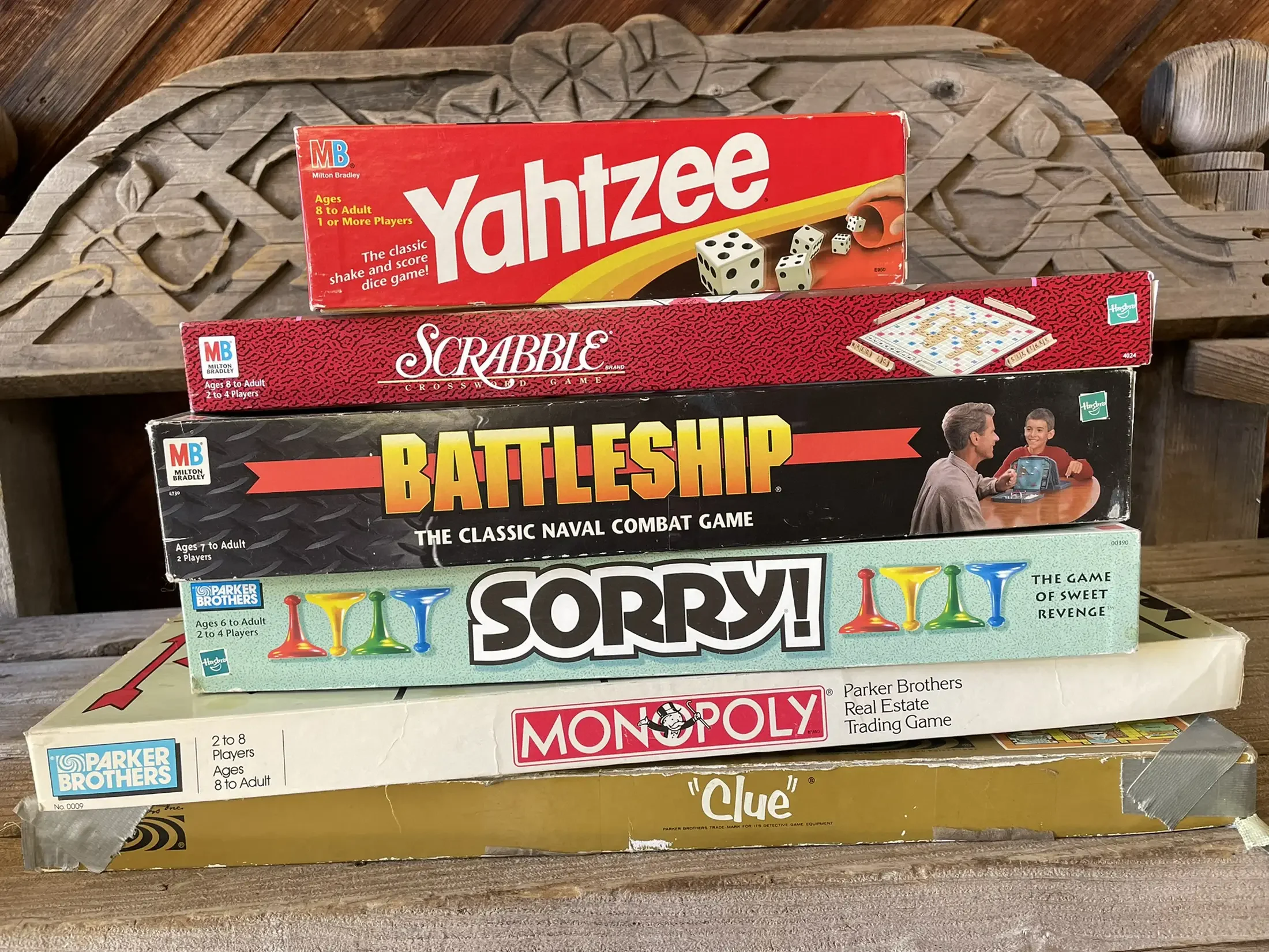 Stack of classic board games including Yahtzee, Scrabble, Battleship, Sorry, and Monopoly on a wooden surface.