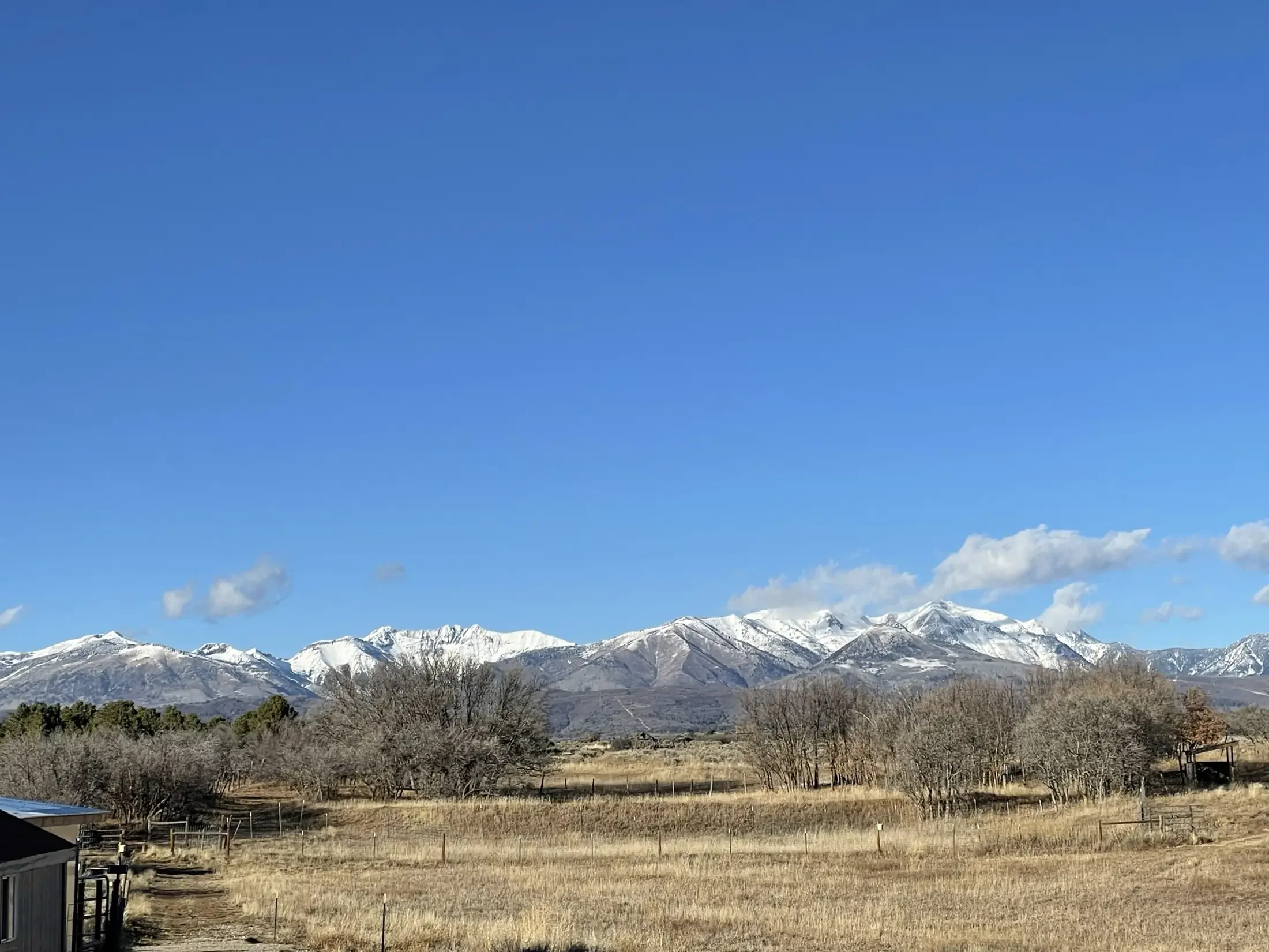 Snow-capped mountains under a clear blue sky, with a rural landscape of trees, fields, and farm buildings in the foreground.