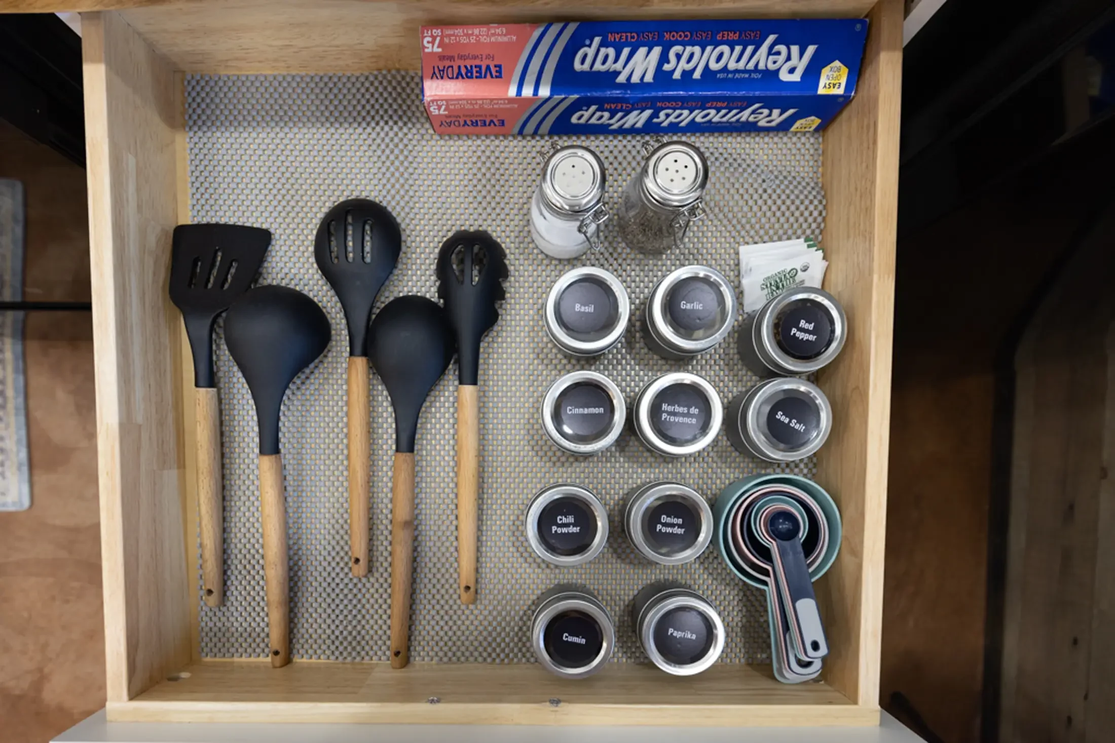 A wooden drawer containing four black kitchen utensils, two shakers for salt and pepper, and several spice jars with labels including basil, garlic, red pepper, cinnamon, herbs de Provence, sea salt, chili powder, onion powder, and paprika. A box of 
