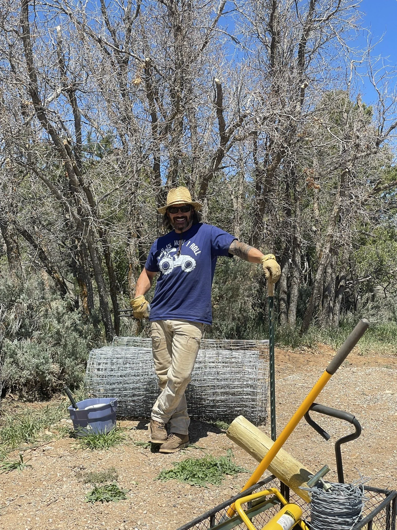 A person wearing a wide-brimmed straw hat, sunglasses, gloves, a blue t-shirt, and khaki pants standing outdoors near a wire cage, gardening tools, and a small bucket, with leafless trees and a clear blue sky in the background.