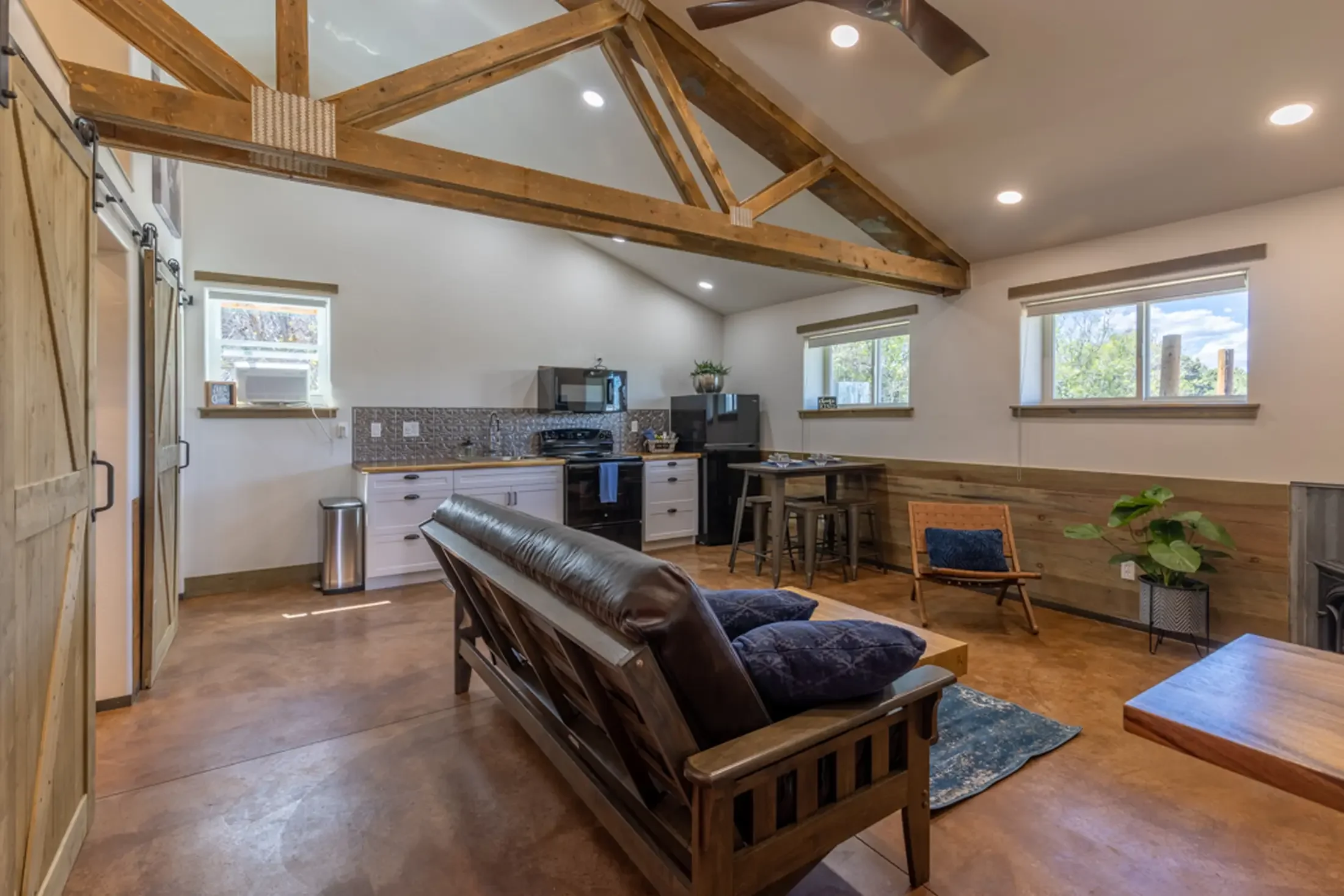 Inside a modern living space with a kitchen and seating area, featuring wooden beams on the ceiling, white walls, hardwood floors, windows with shades, and furniture including a leather armchair and wooden table.