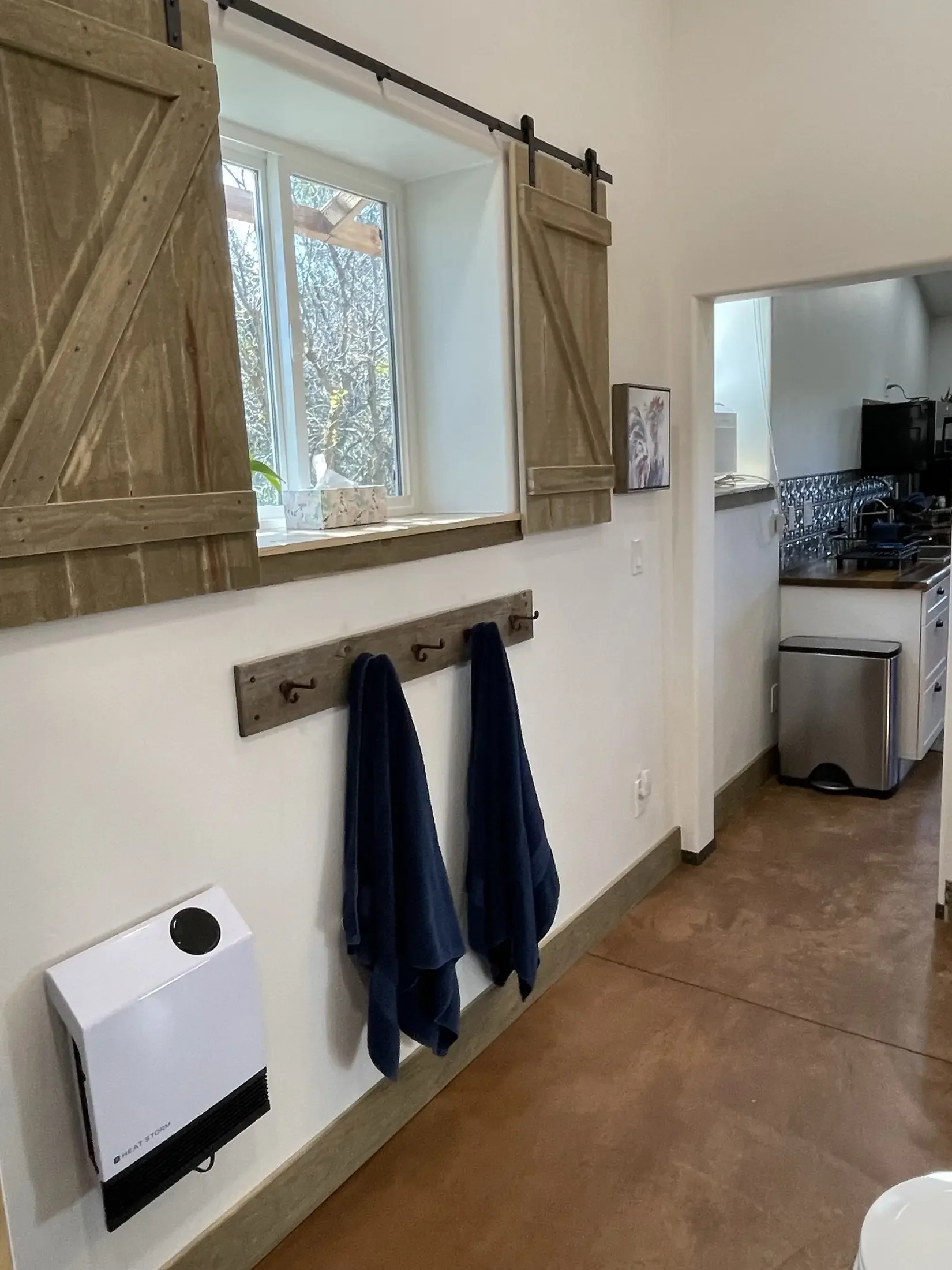 Interior view of a room with a window, wooden sliding barn doors, hooks with dark clothing, and a wall-mounted heater.