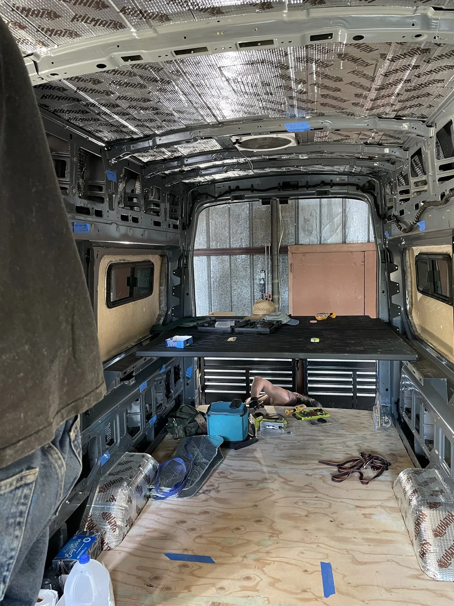 The interior of a vehicle being renovated or built. The ceiling and walls are lined with reflective insulating material. The floor is covered with plywood, and various tools and supplies are scattered around, including a blue toolbox, wire, a water j