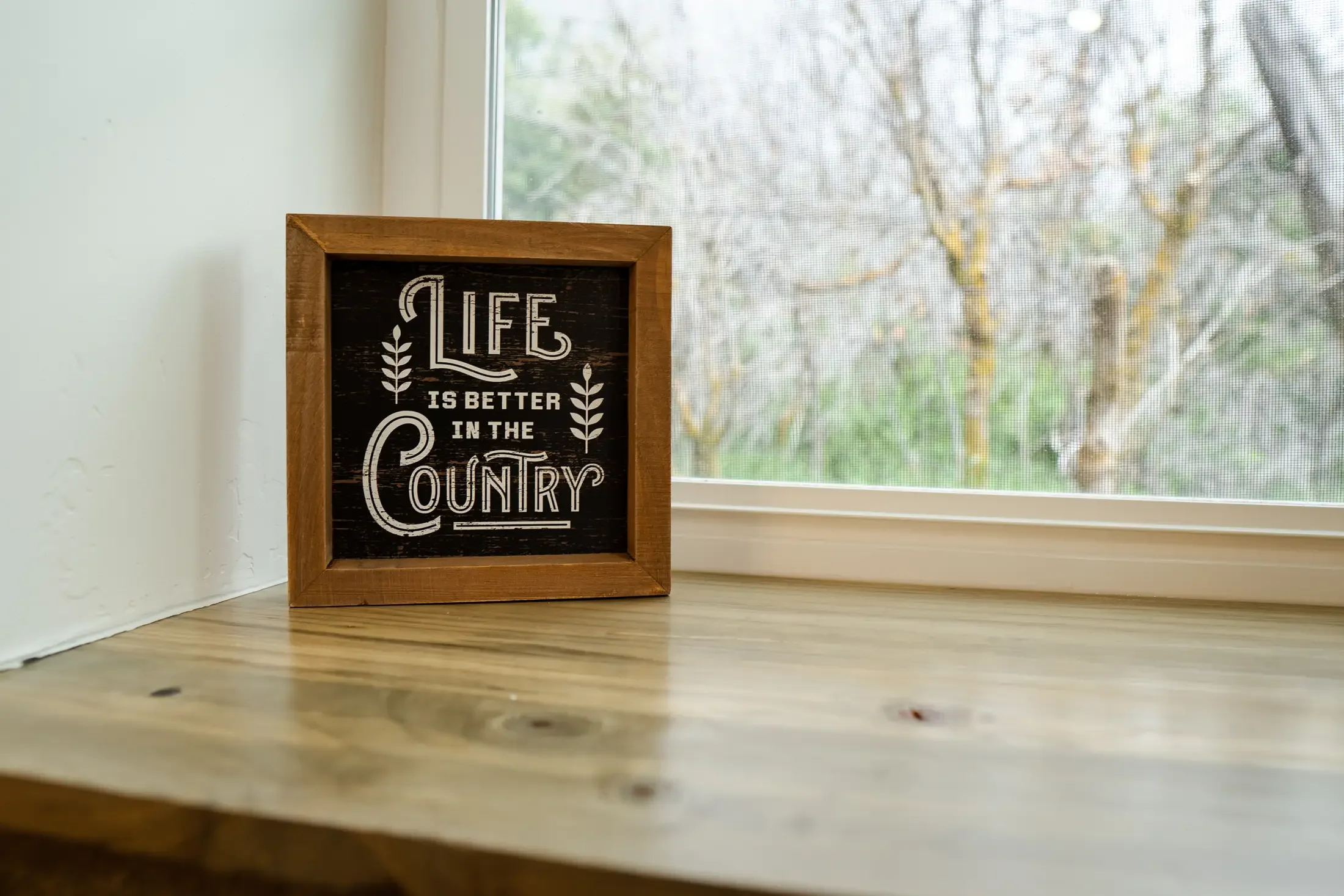 A small wooden framed chalkboard with the words 'Life is better in the country' written in white, placed on a wooden surface near a window with a view of bare trees outside.