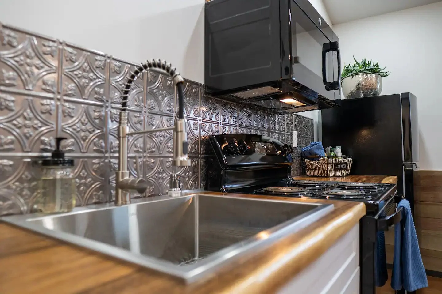 Modern kitchen with stainless steel sink, black microwave and stove, decorative metal tile backsplash, potted plant on black cabinet, and wooden countertop.