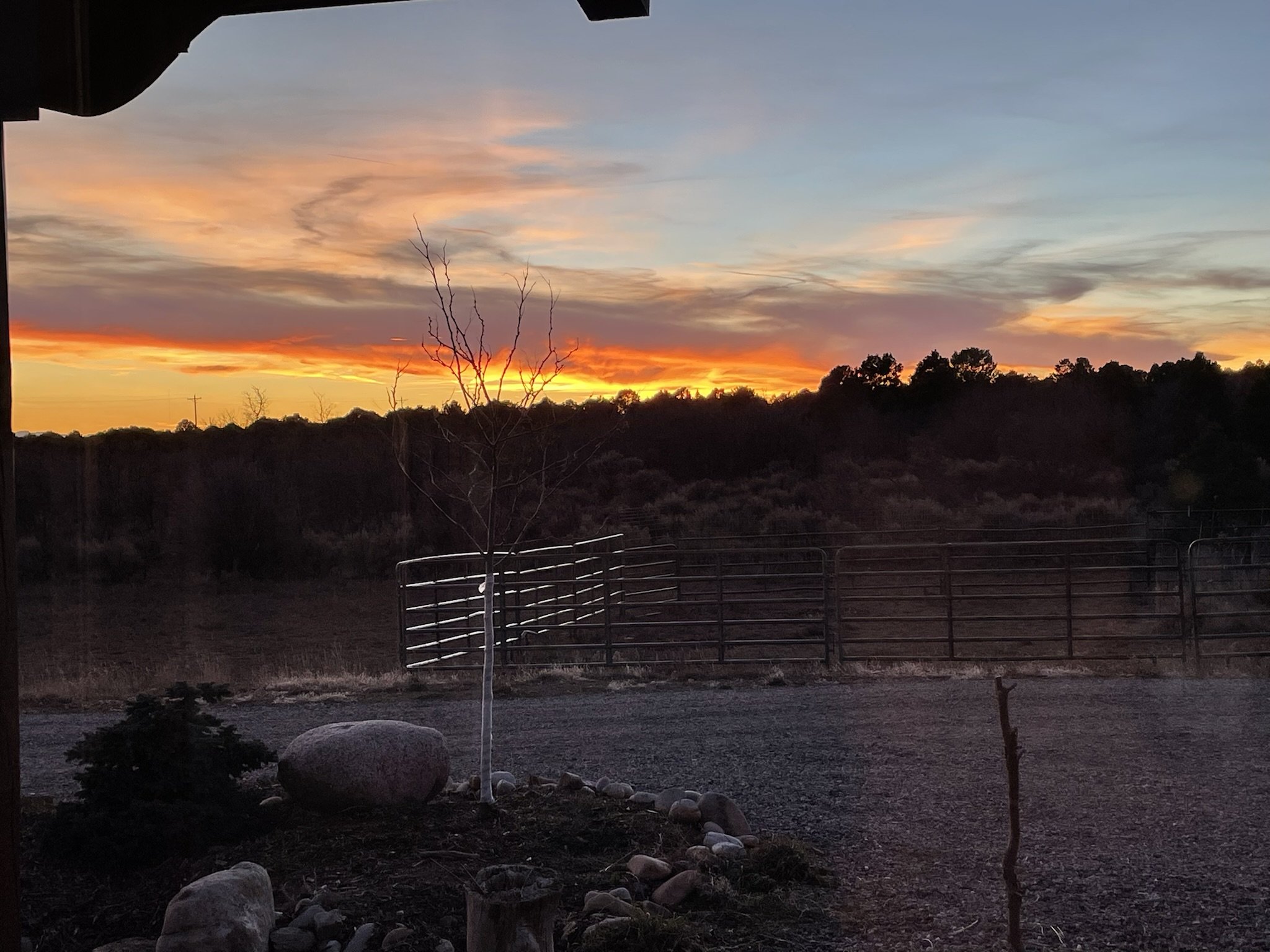 A sunset with colorful clouds over a landscape with trees and fences, seen from a porch.
