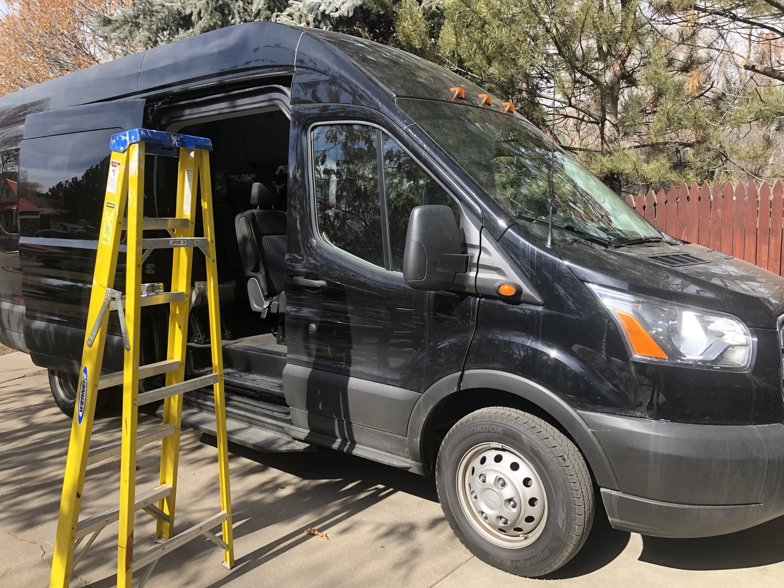 A black van parked outdoors with a yellow ladder leaning against its side. The van is surrounded by trees with autumn foliage, a wooden fence, and sunlight reflected on its surface.