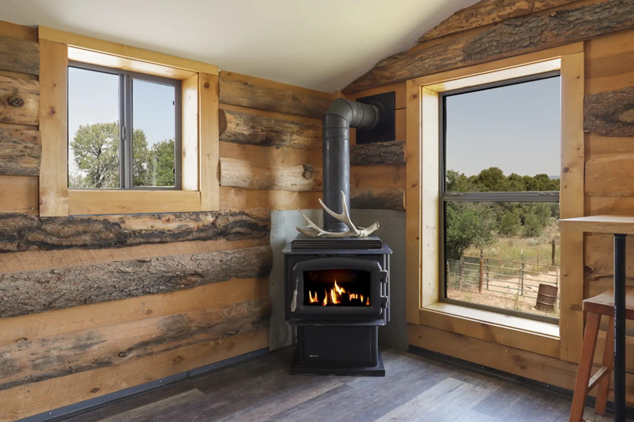 Interior of a log cabin with two windows showing trees outside, a wood stove with a fire, and a deer antler on top.