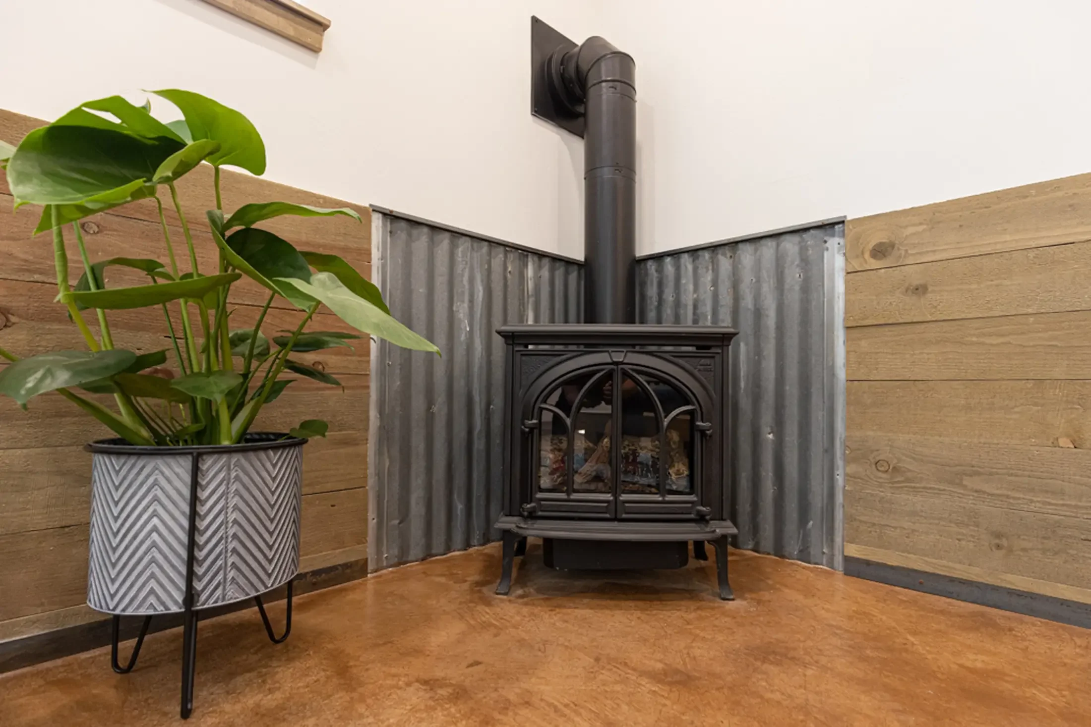 A potted plant with green leaves on a metal and wood stand, with a wood and metal wall and a wood stove in the background.