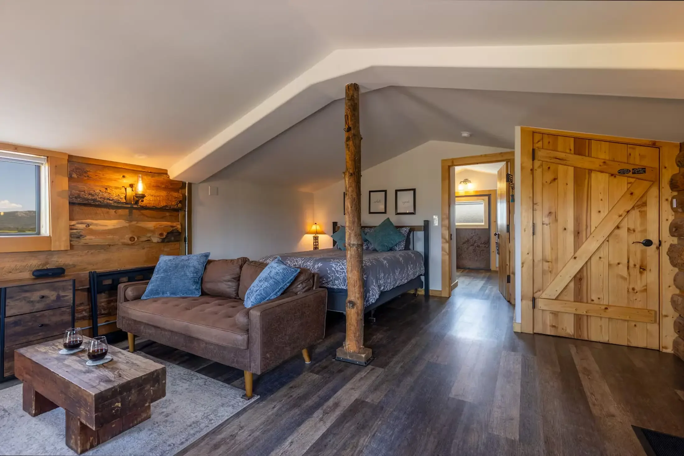 Interior of a rustic bedroom and living area with wooden accents, a corner bed, a leather sofa with blue pillows, a wooden coffee table with wine glasses, and a wooden door, with a large window showing mountains outside.