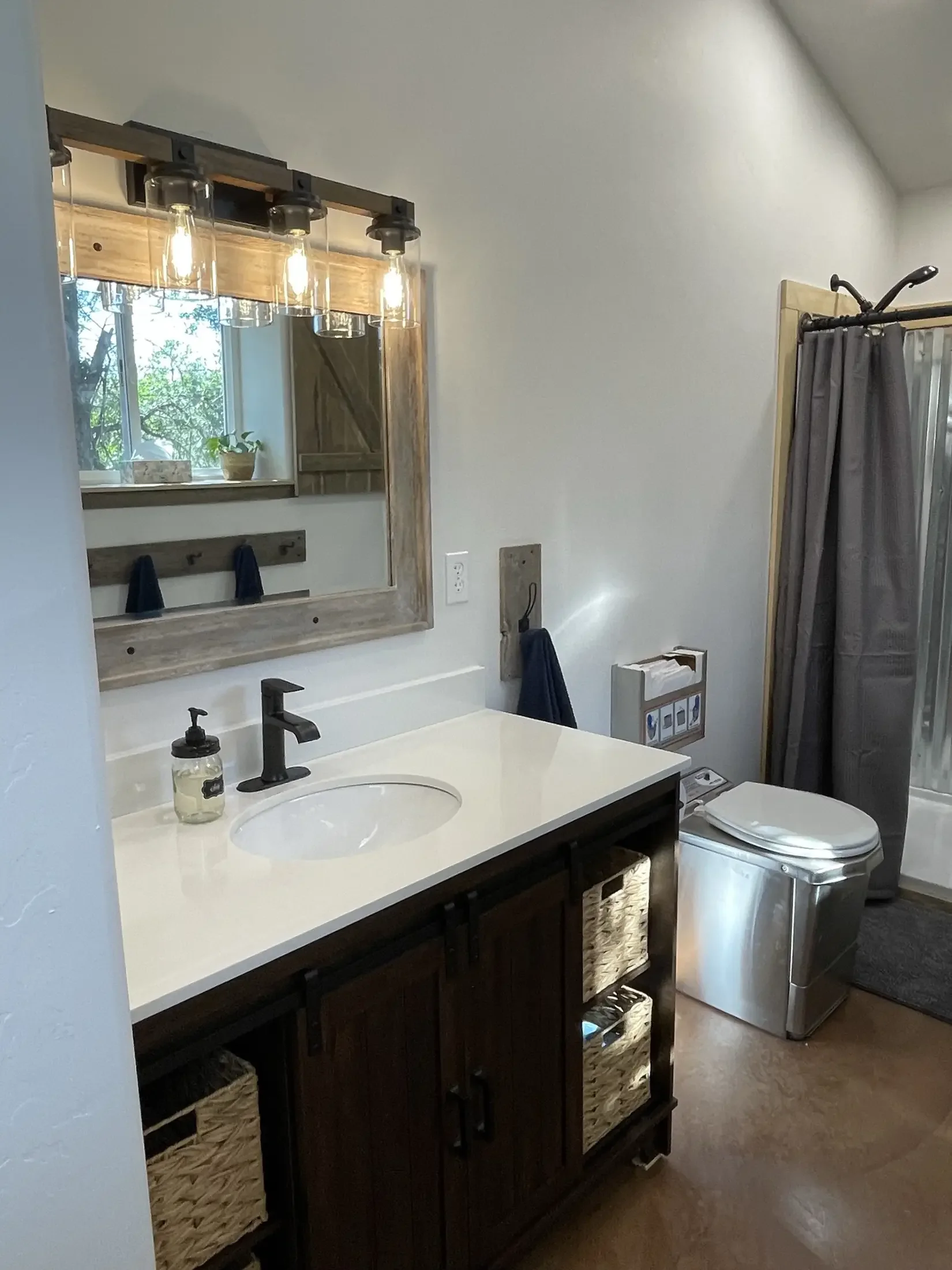 Bathroom vanity with dark wood cabinets, white countertop, and a black faucet. A soap dispenser is on the counter. Above the sink, a mirror is framed with wood, with a window reflected in it showing outdoor greenery. There is a light fixture with mul