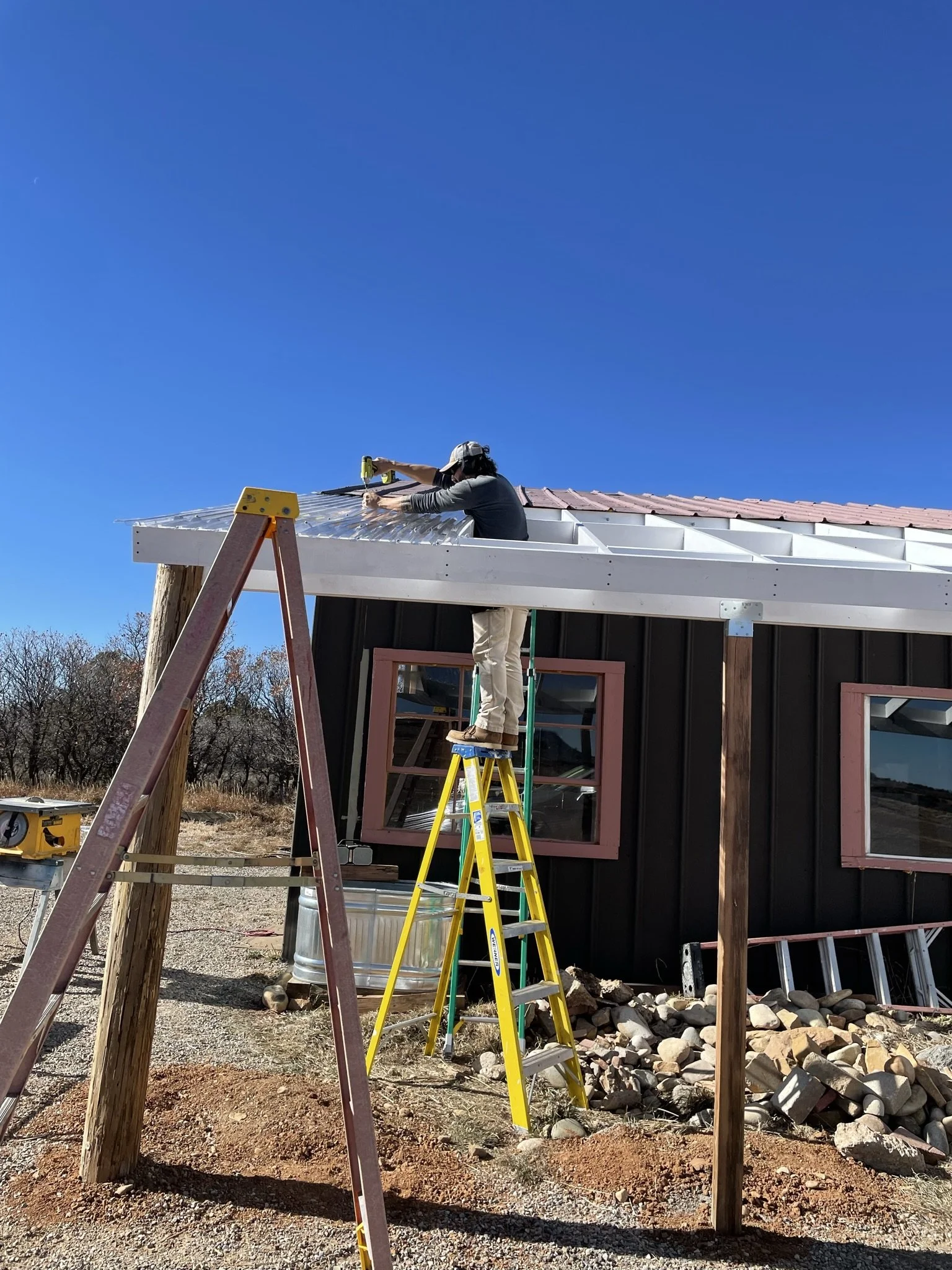 A person is working on a roof, standing on a ladder and using a power drill to install metal roofing panels on a building under construction. The building has black walls with pink window frames, and the sky is clear and blue.